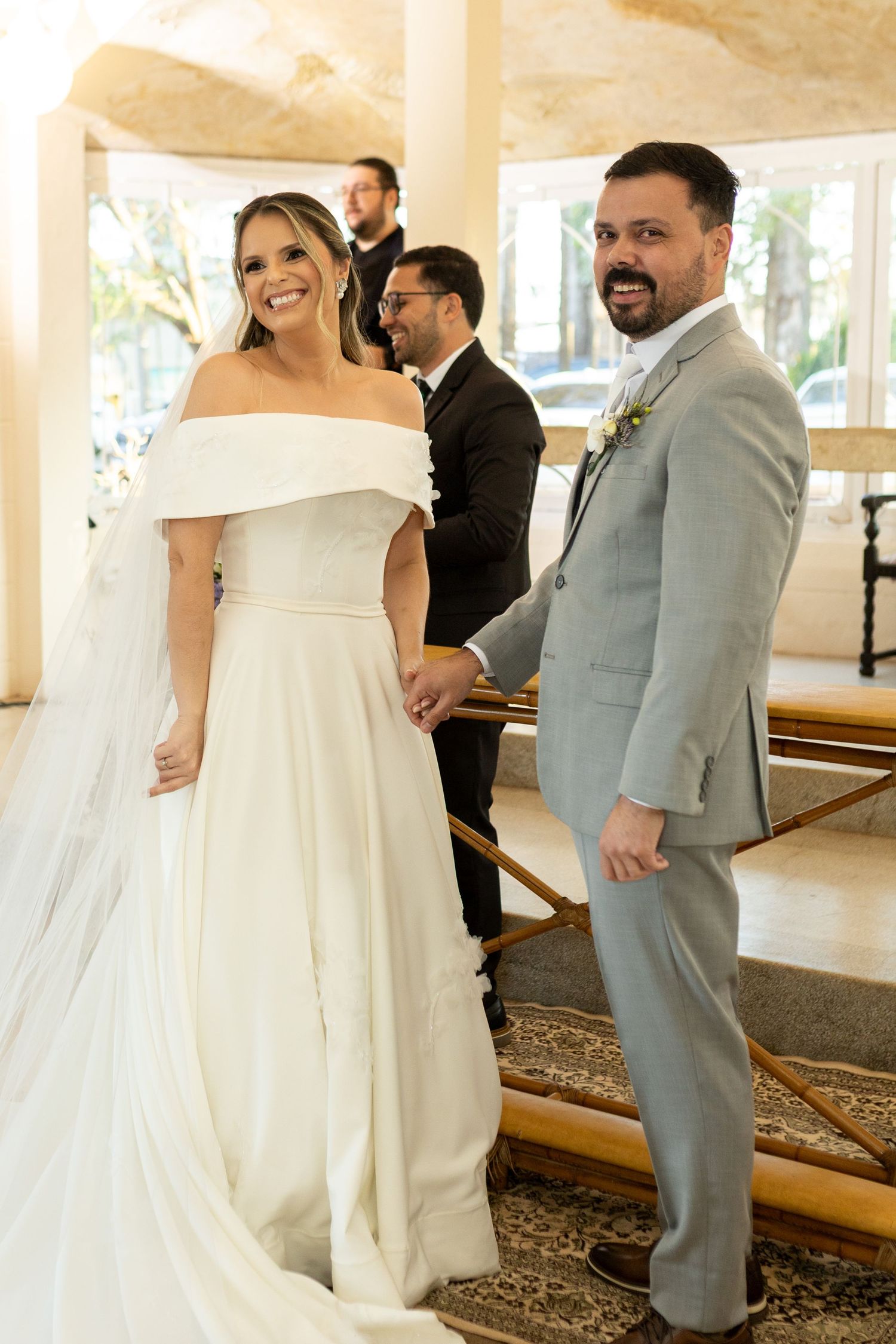 Smiling bride in off-shoulder white gown holds hands with groom in gray suit at elegant wedding ceremony.