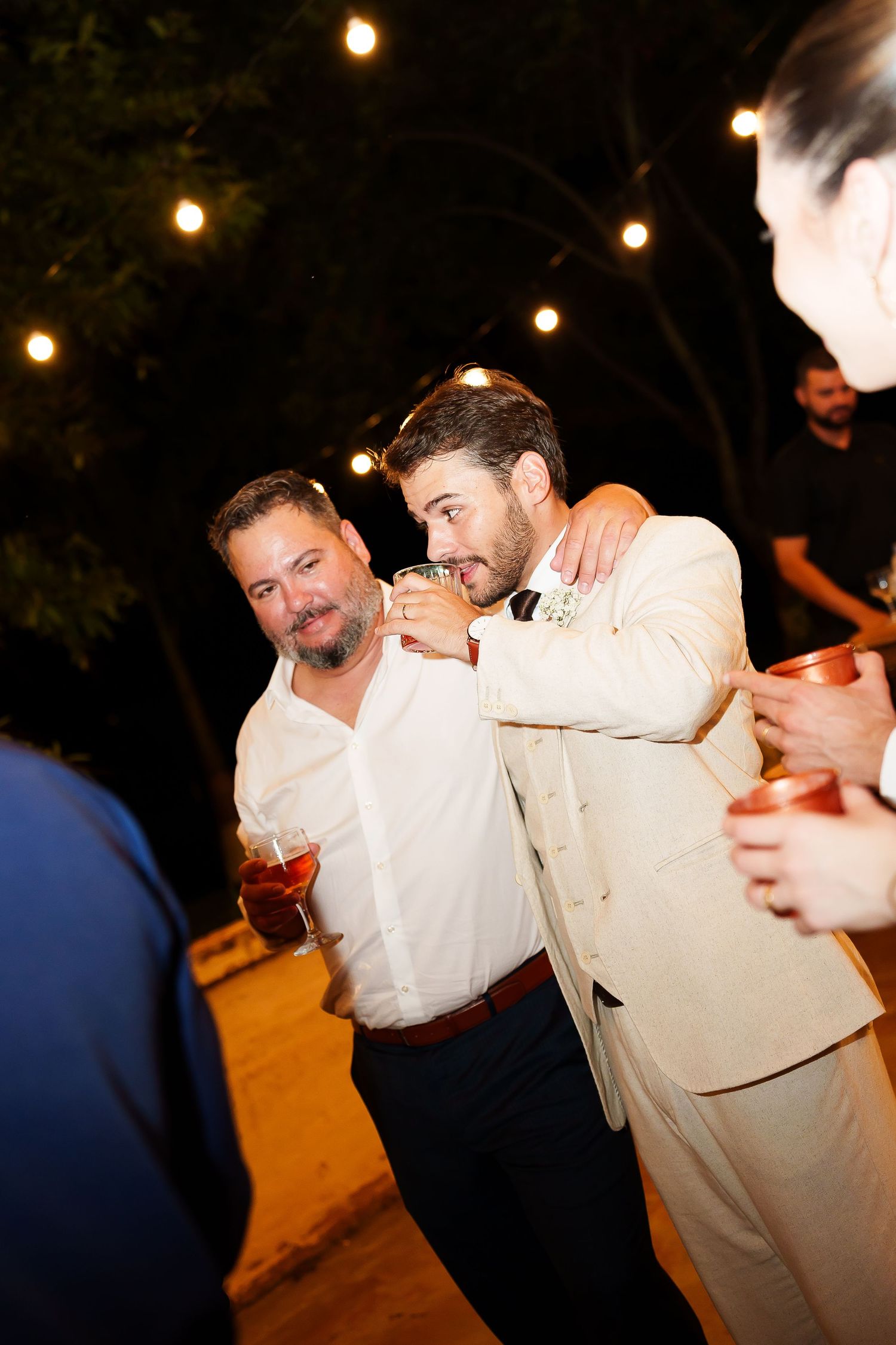 Two men in formal attire share a friendly embrace at an outdoor evening wedding reception with string lights.