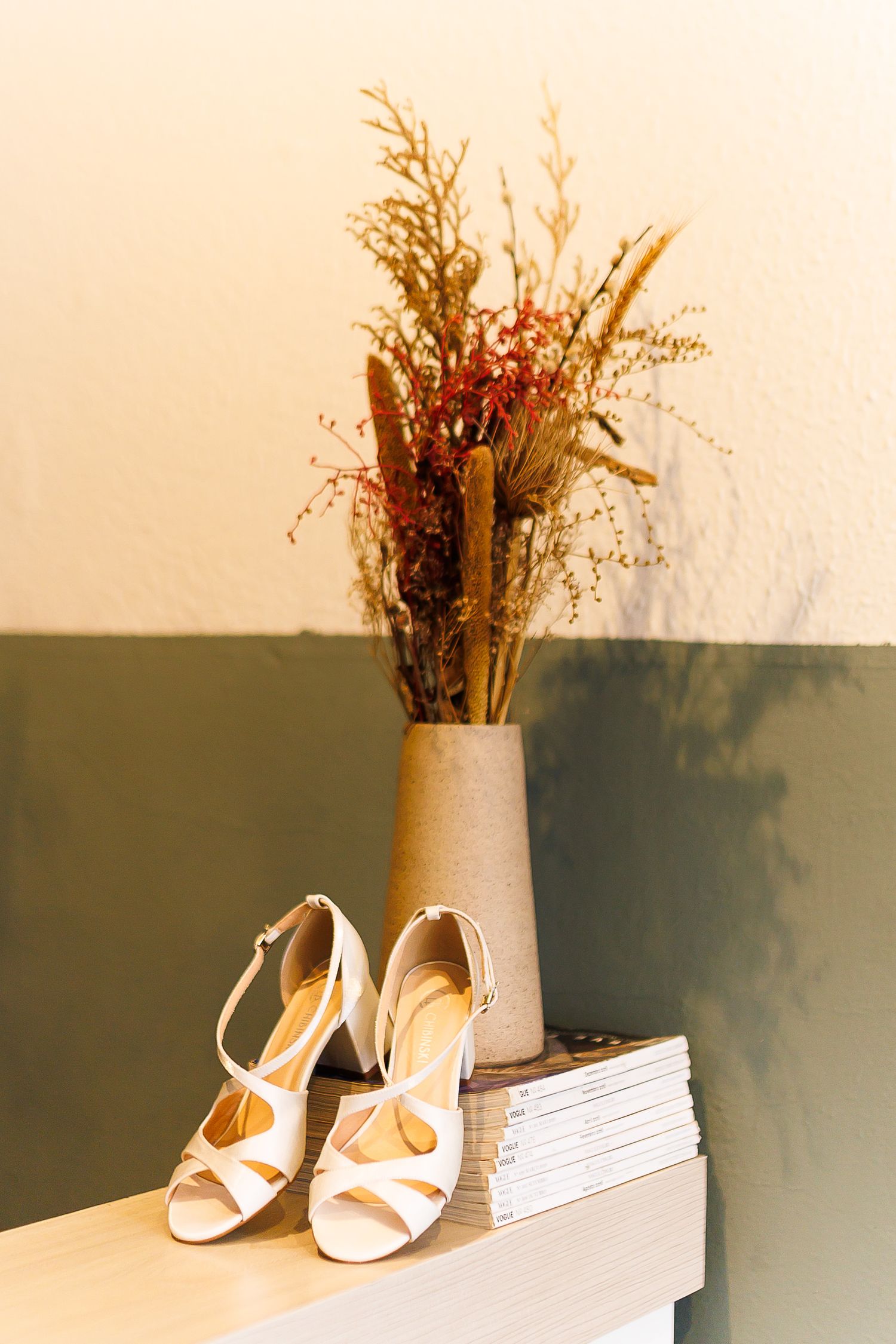 White strappy heeled sandals on stacked magazines beside a rustic vase with dried botanicals on a shelf.