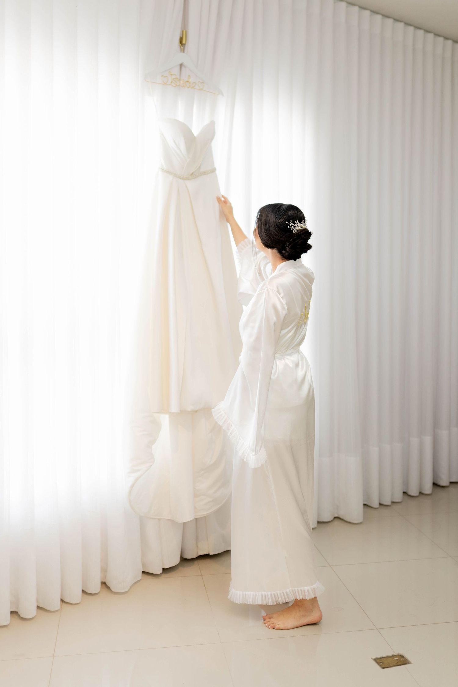 Bride in white robe reaching for her wedding dress hanging by a bright window on her wedding morning.