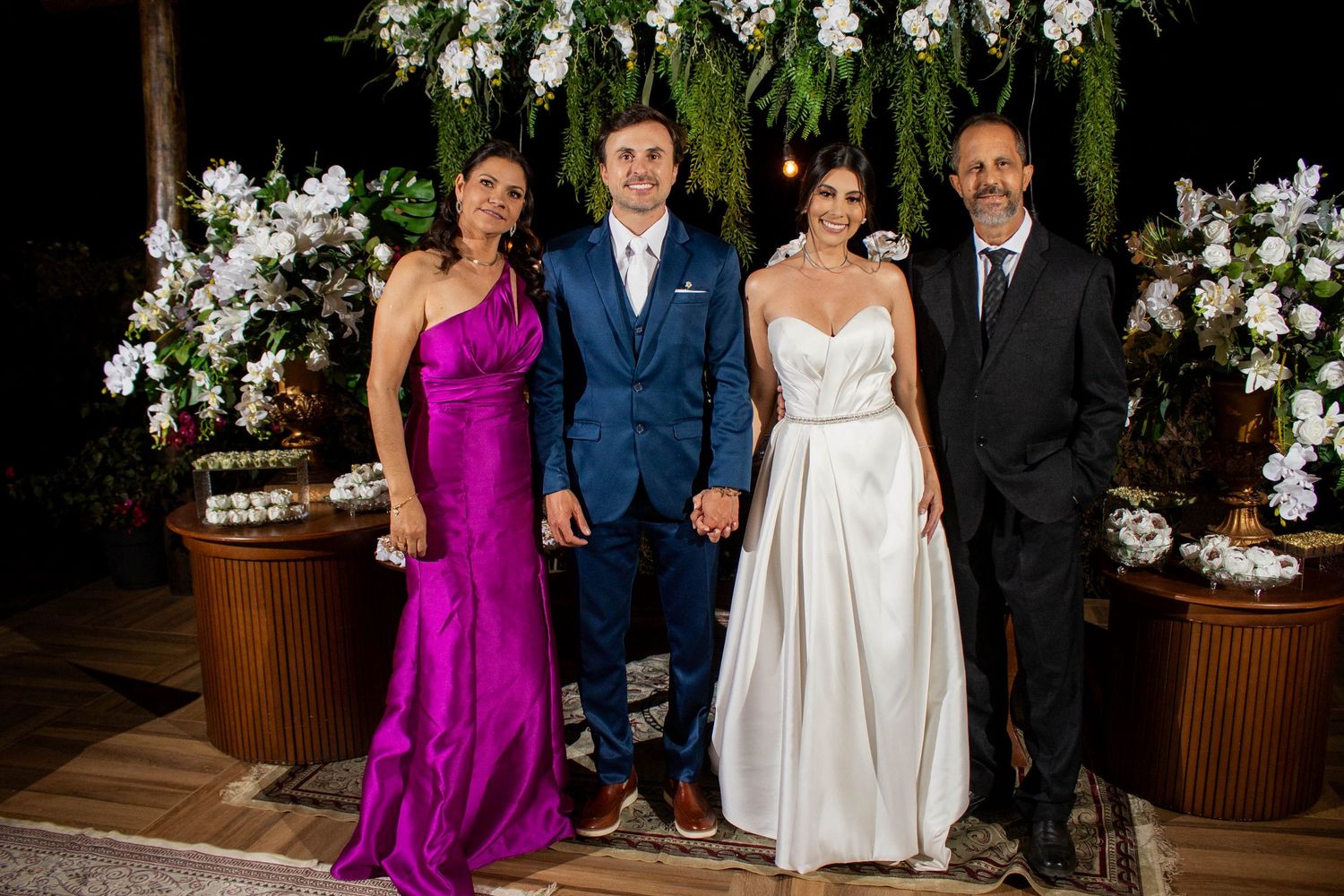 Wedding party of four posing together, bride in white gown, groom in navy suit, woman in magenta dress, man in black suit.