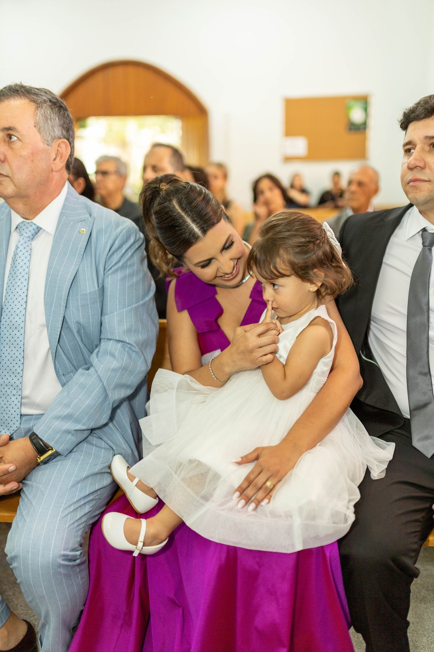 Woman in purple dress holds toddler girl in white dress at a formal church ceremony with guests seated in pews.