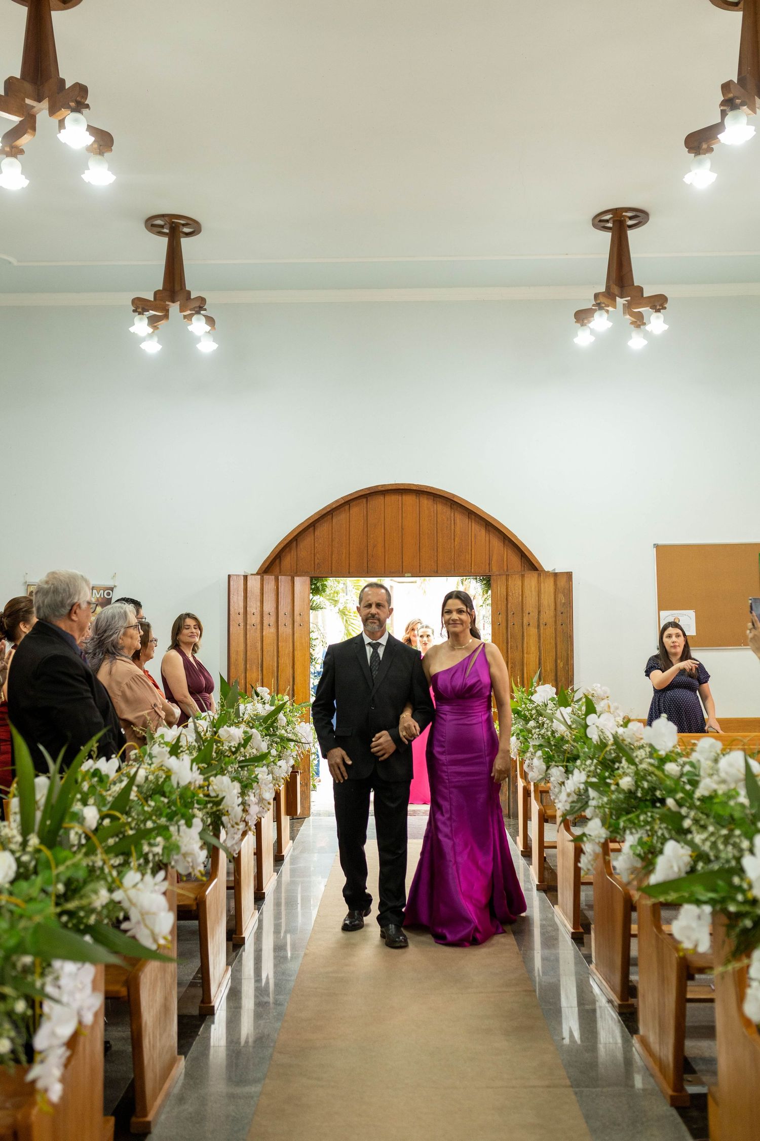 A couple walks down a flower-adorned church aisle, the woman in a purple gown, during a wedding ceremony.