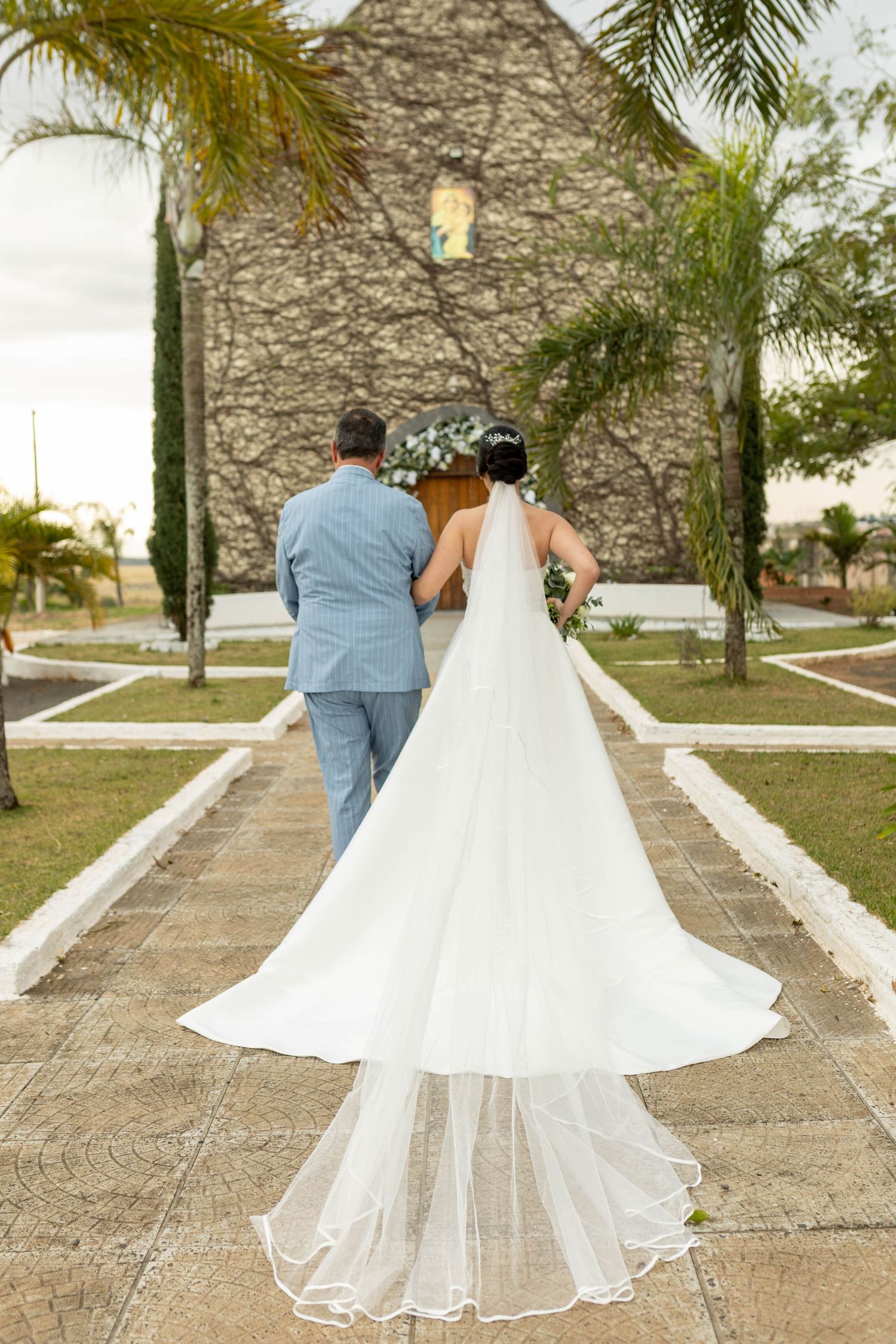 Bride in white gown with long veil and groom in blue suit walk toward stone chapel surrounded by palm trees.
