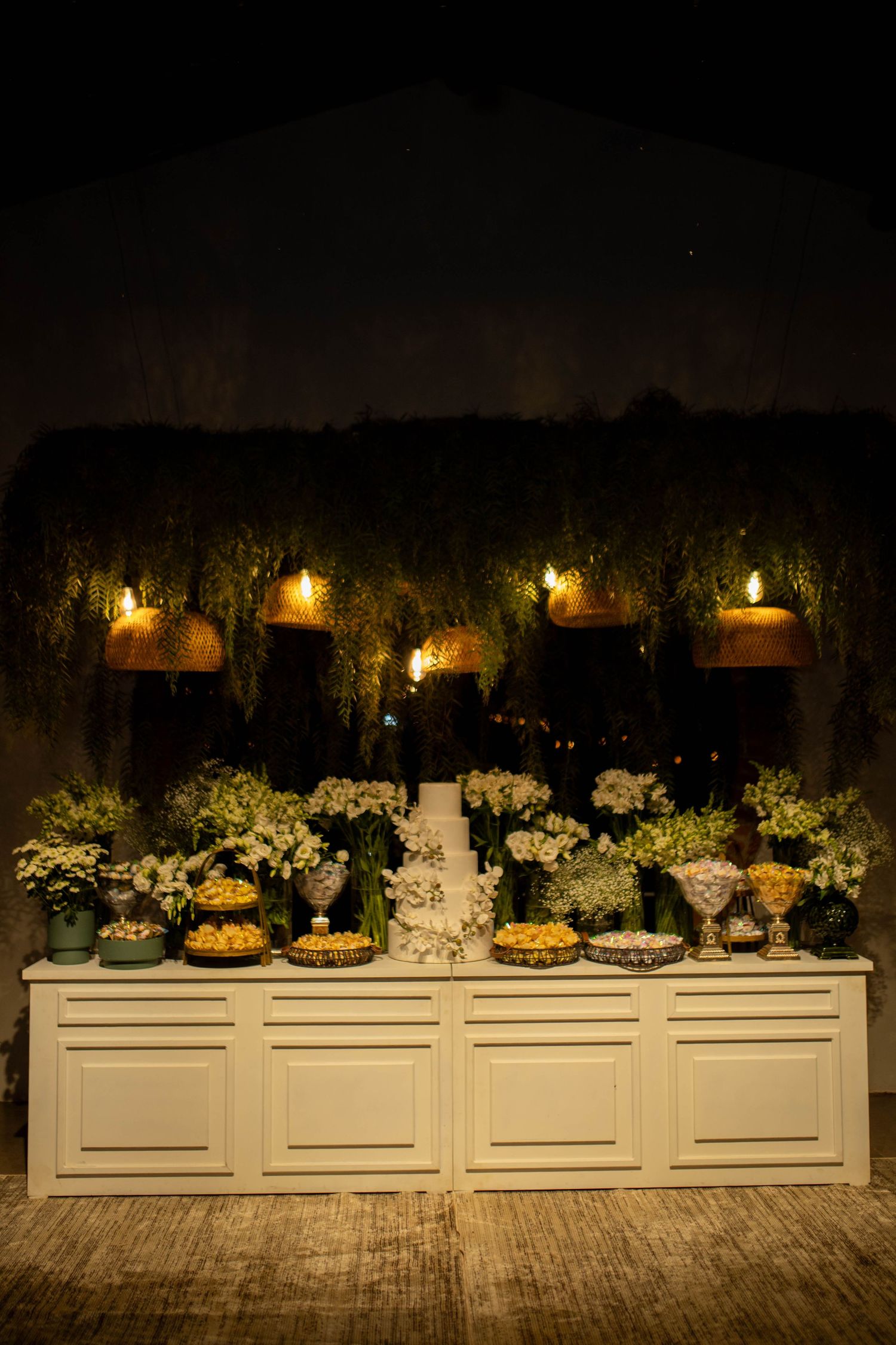 Elegant wedding dessert table with white flowers, candles, and copper pendant lights in a romantic nighttime setting.