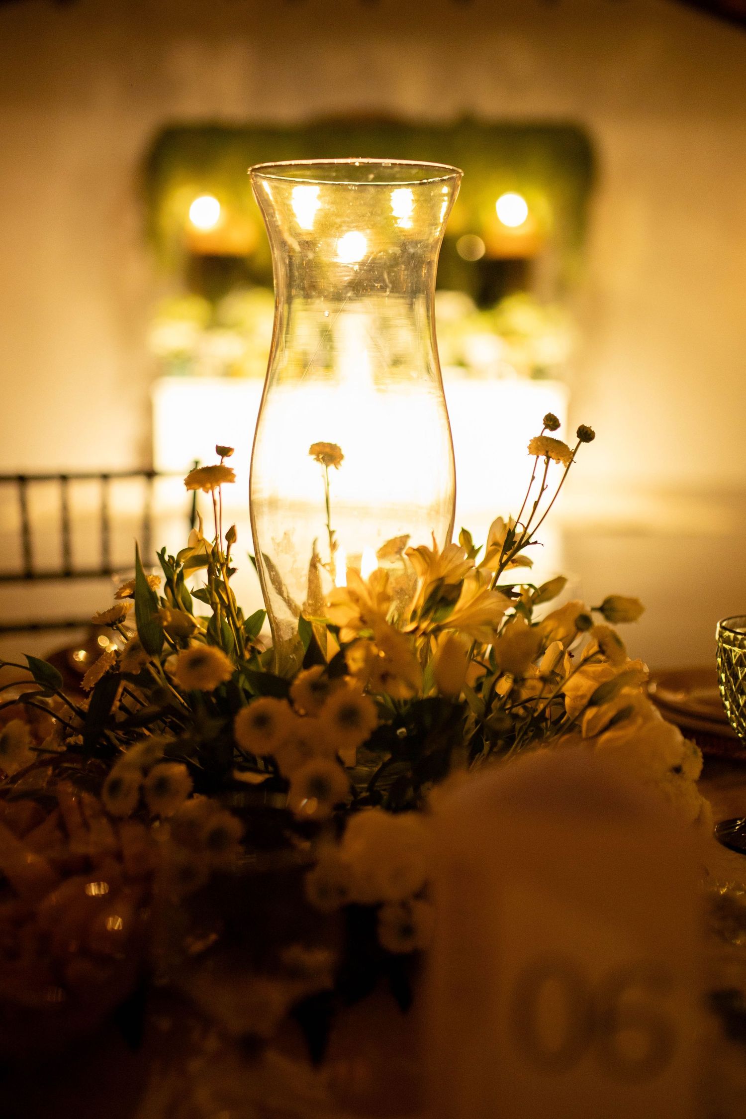Elegant wedding table centerpiece with glass hurricane candle holder surrounded by white flowers in warm golden light.