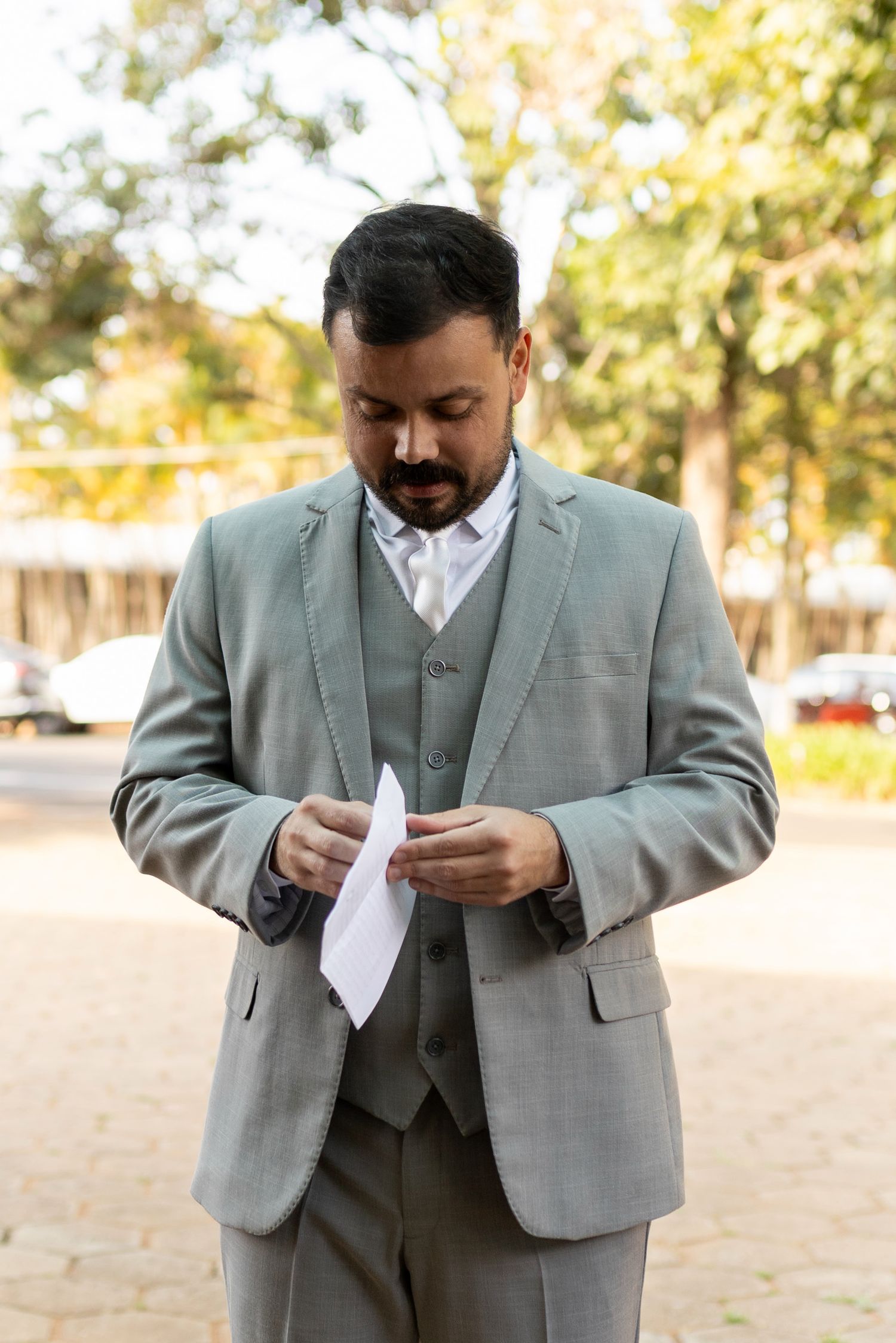 Bearded man in gray three-piece suit looking down at paper outdoors.