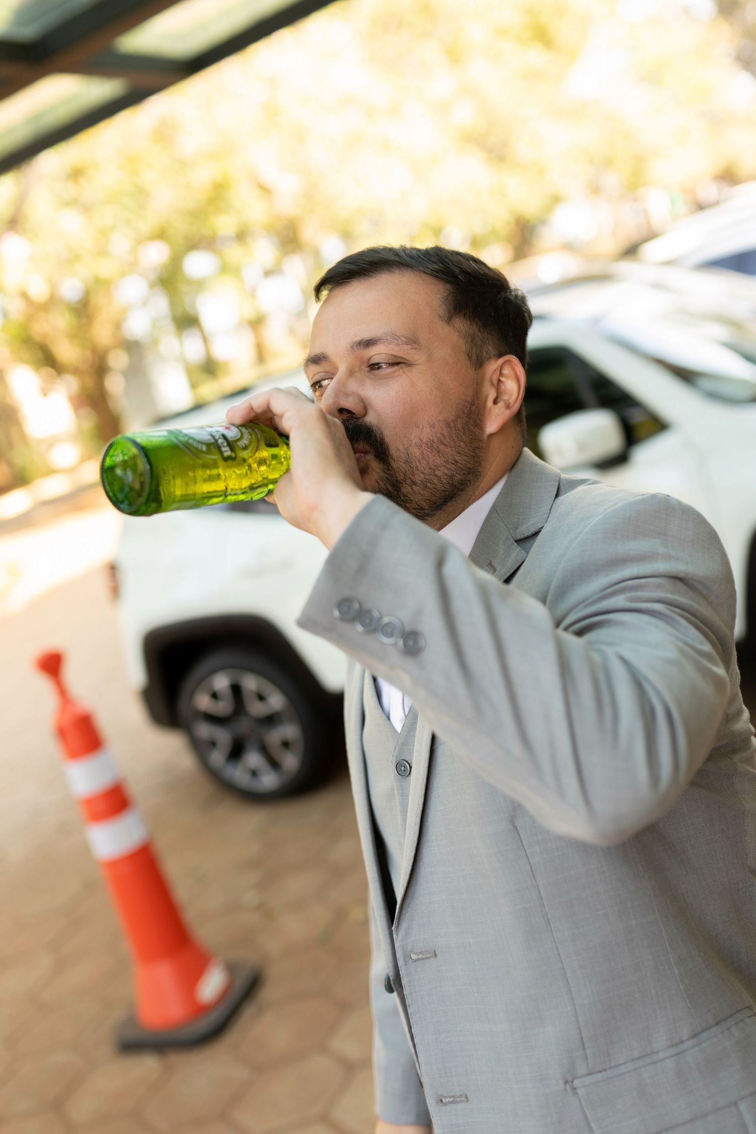 A businessman in a gray suit drinks from a green bottle outdoors near a white car and orange traffic cone.