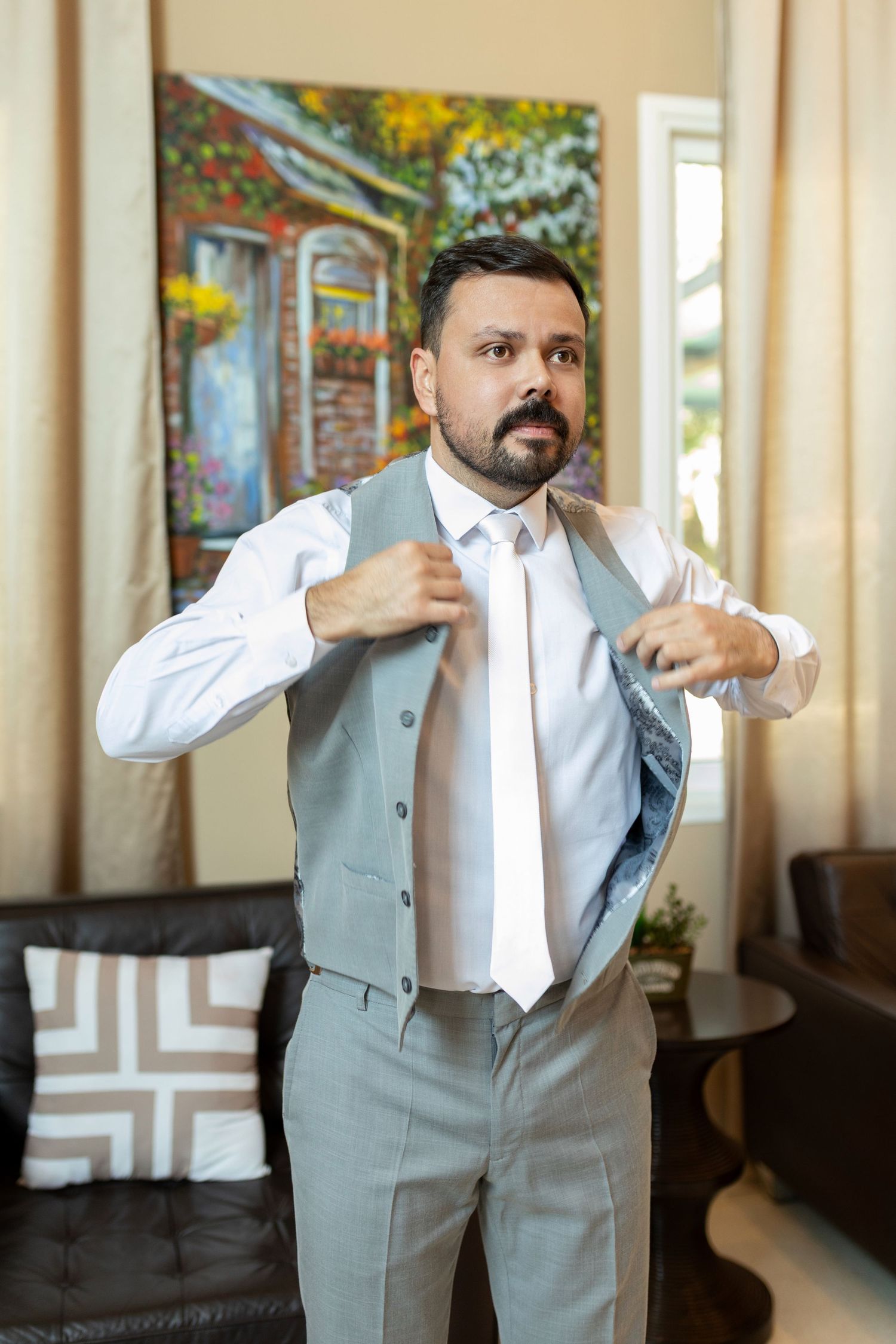 Bearded man in gray vest and white dress shirt adjusting his suit jacket indoors.