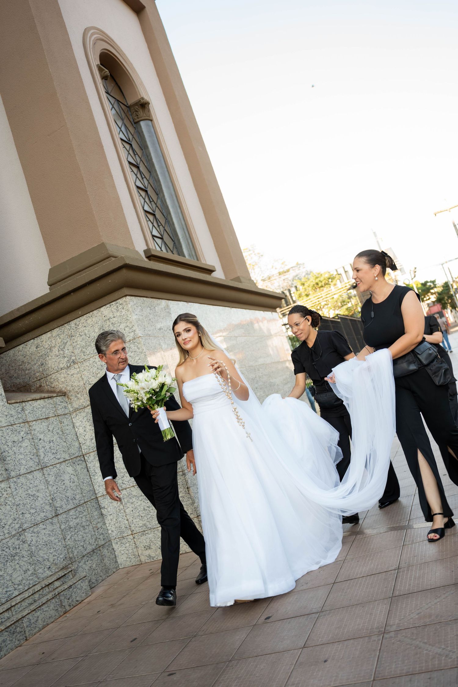 Bride in white gown walks with wedding party outside church, attendants carrying her train.