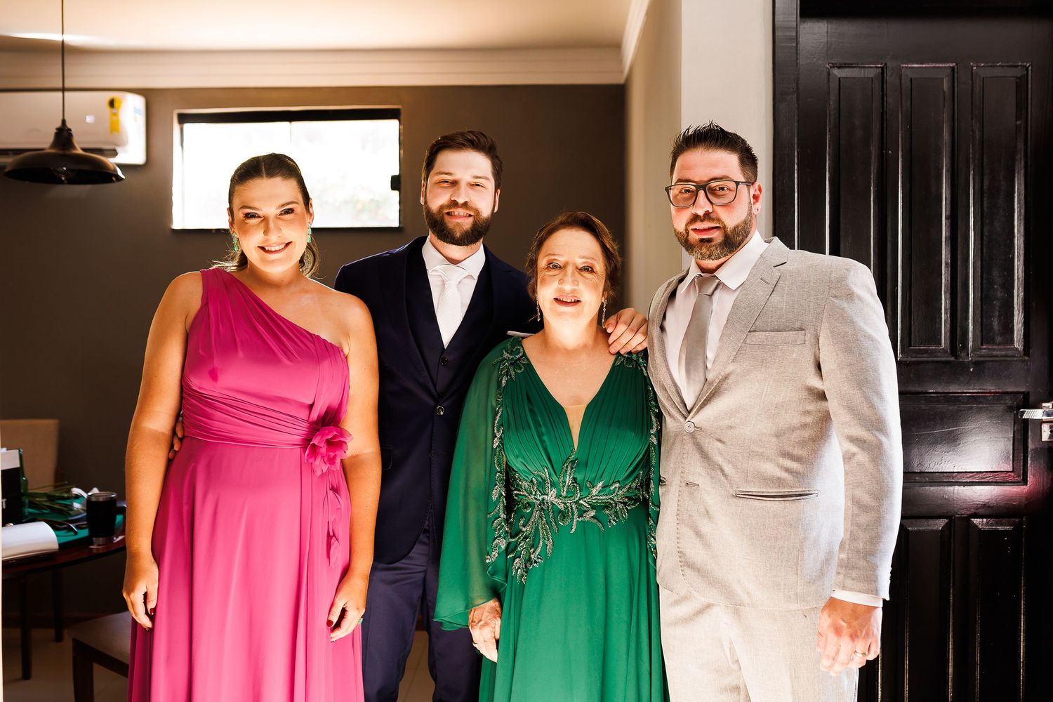 Four elegantly dressed people posing together indoors, wearing formal attire including pink and green gowns and suits.