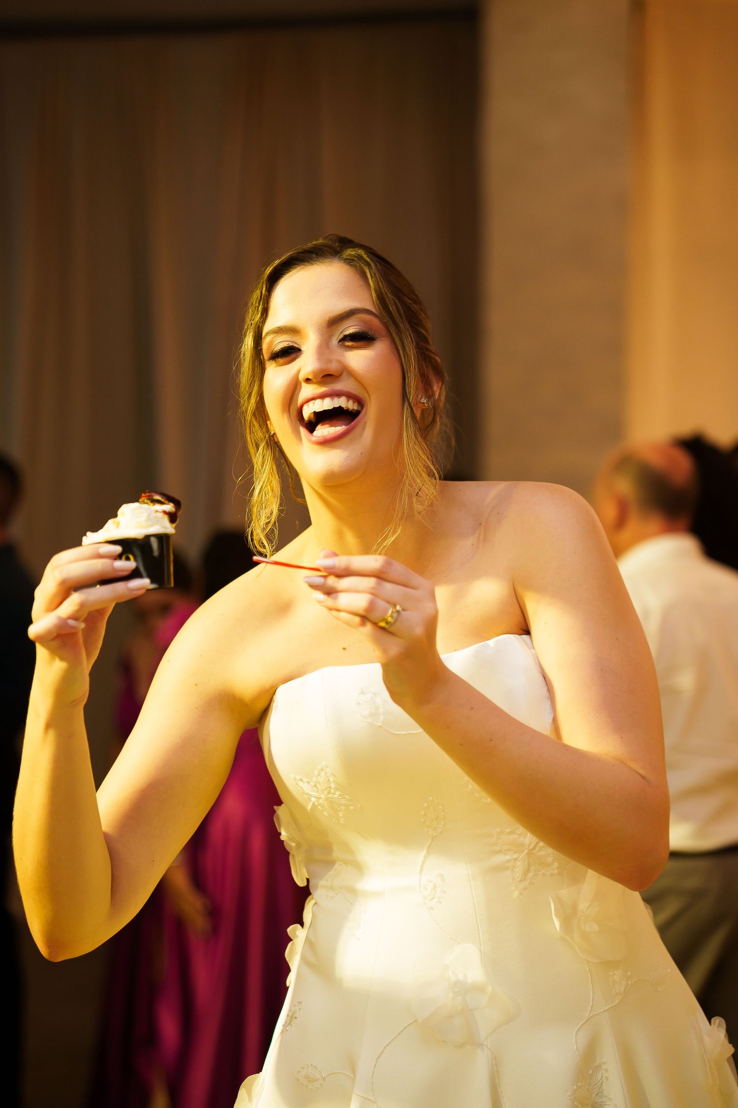 Laughing bride in strapless white gown holding a cupcake at her wedding reception.