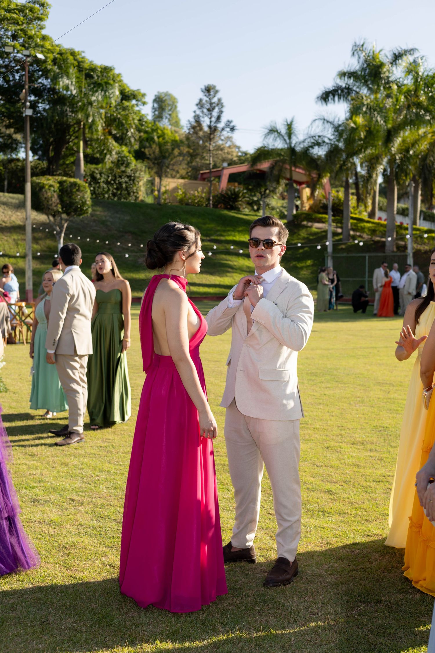 Couple in formal attire at outdoor wedding, woman in pink gown, man in white suit adjusting bow tie on green lawn.