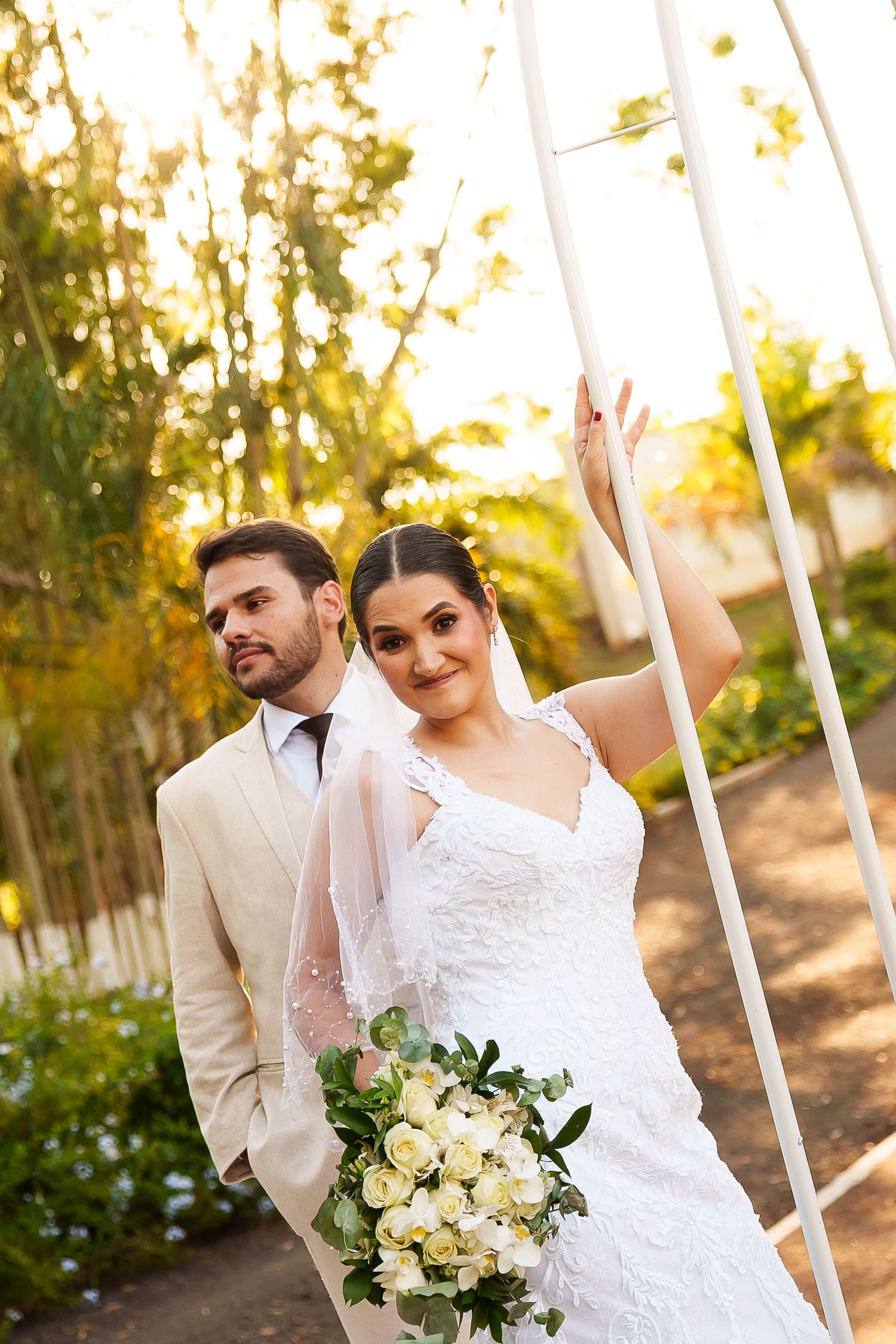 Bride holding white roses bouquet and groom in beige suit posing outdoors in a garden during golden hour.