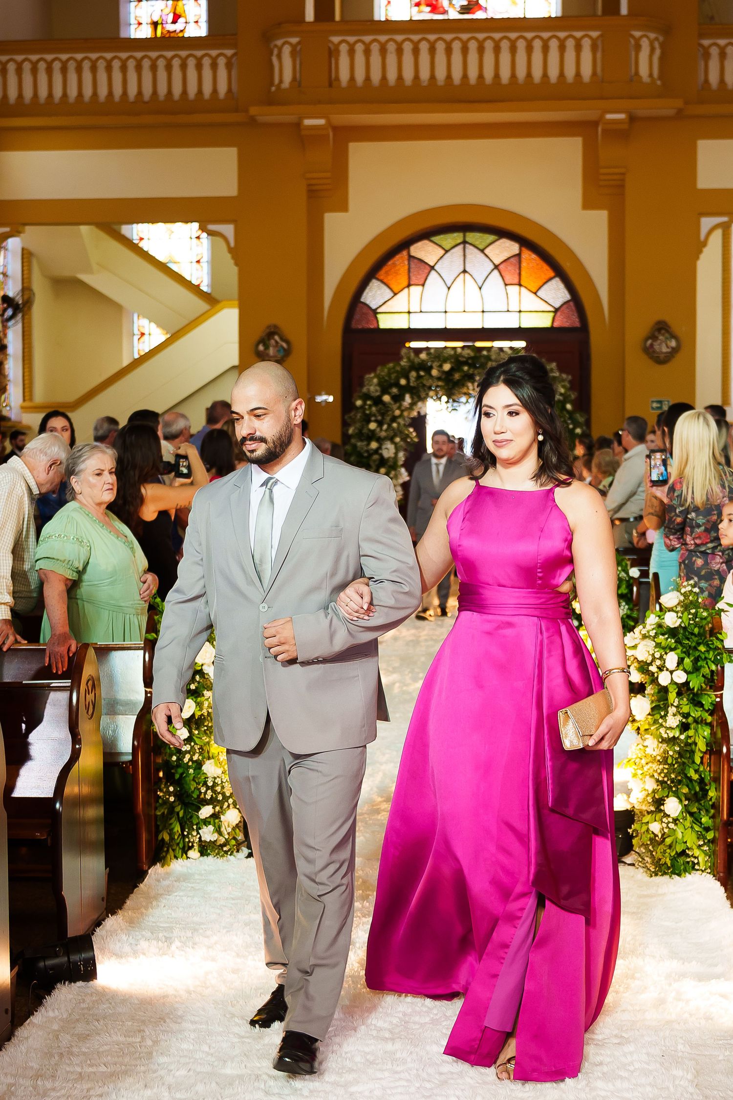 Couple walking down church aisle, man in gray suit, woman in pink gown, floral decorations.