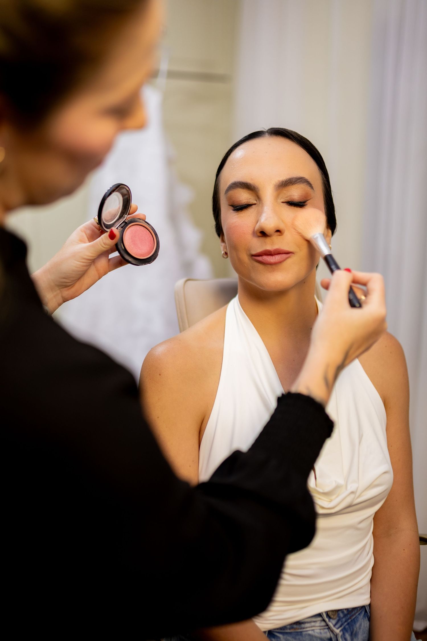 A makeup artist applying blush to a young woman's face with a brush in a bright beauty studio.