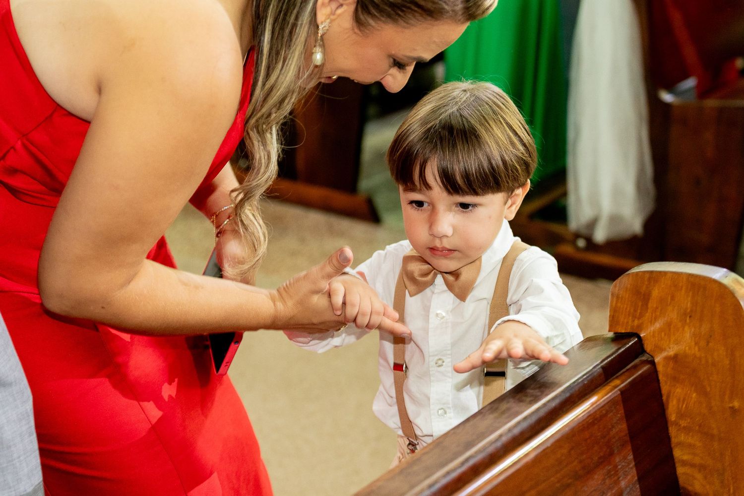 A woman in a red dress interacts with a young boy in suspenders and bow tie at a church pew.