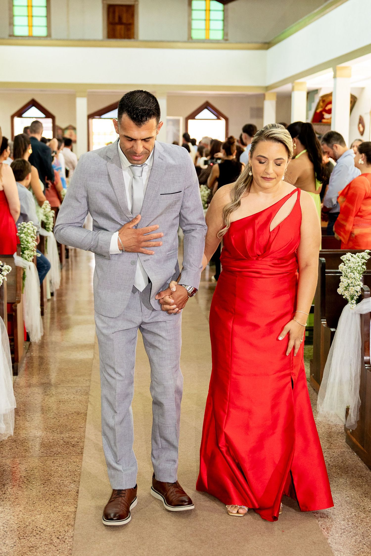 A man in a gray suit escorts a woman in a red one-shoulder gown down a church aisle at a wedding.