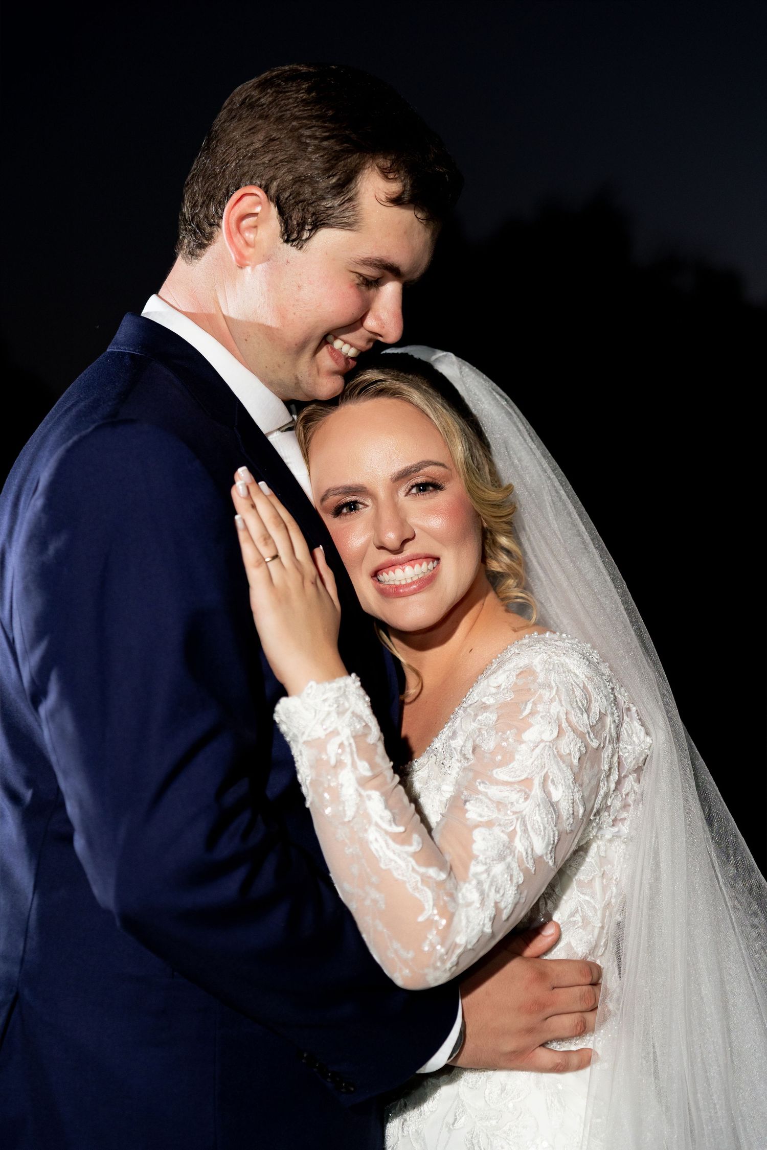 Bride in lace gown and veil smiles with groom in navy suit during romantic nighttime wedding portrait.
