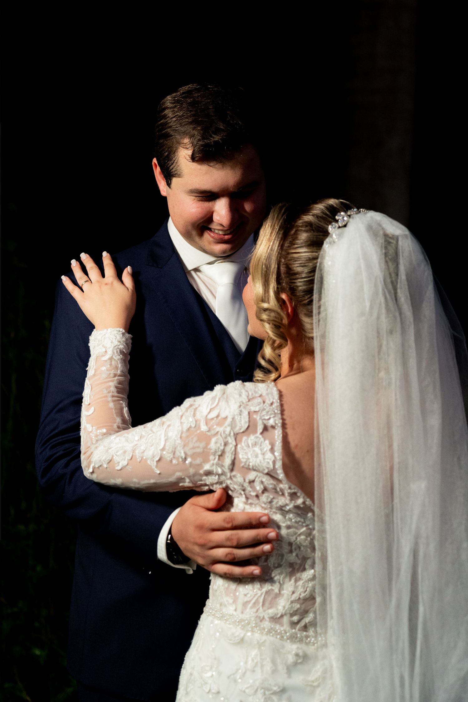 Bride in lace gown and veil embraces smiling groom in navy suit during romantic wedding portrait.
