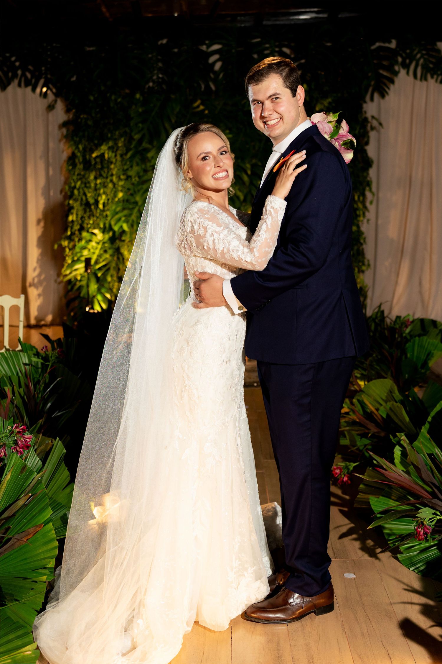 Bride in lace gown and veil poses with groom in navy suit, embracing before lush tropical greenery backdrop.