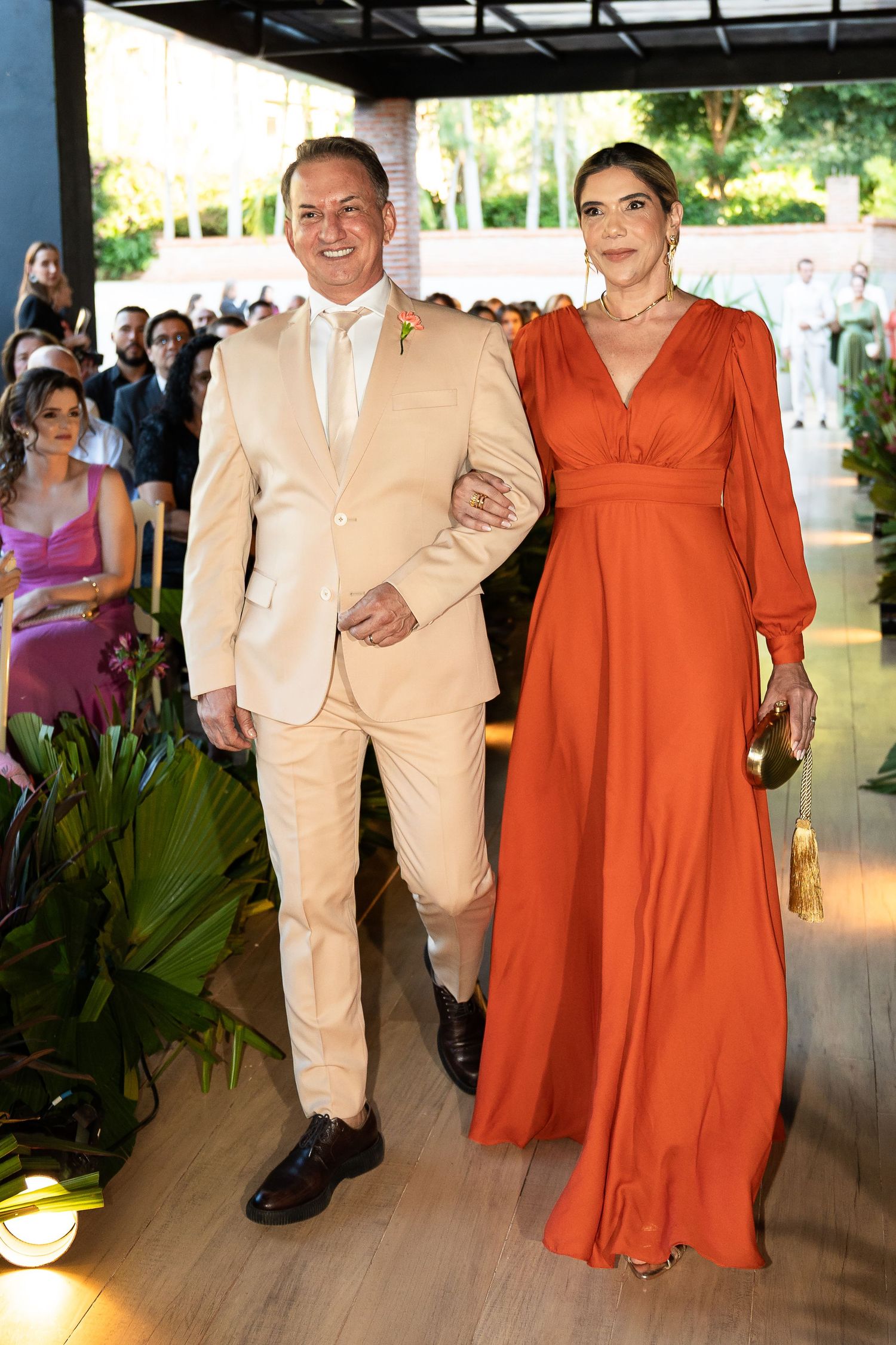 Elegant couple posing at a formal event, man in beige suit, woman in flowing orange gown, lush greenery backdrop.