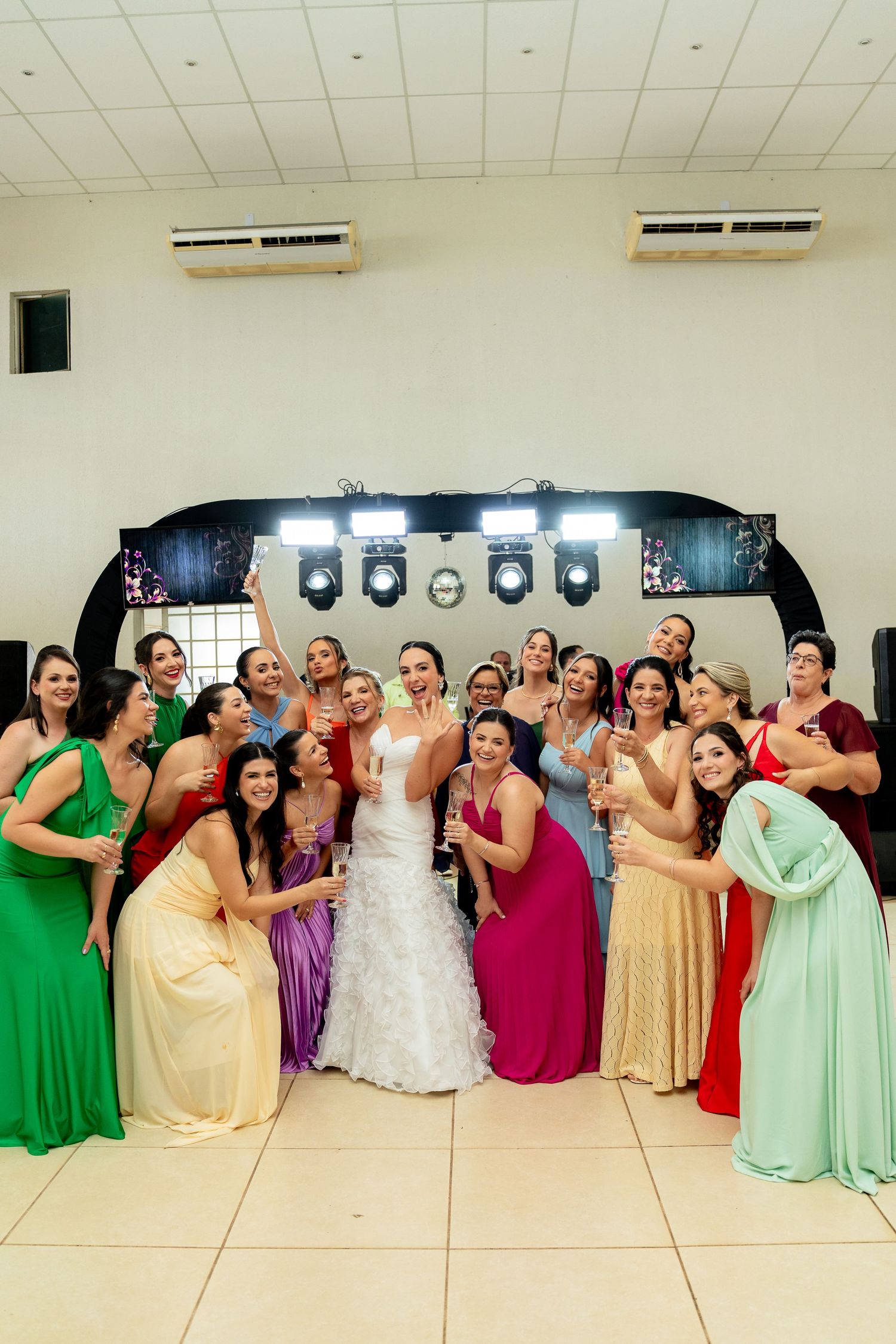 Bride in white gown poses with bridesmaids in colorful dresses, raising glasses at a wedding reception.