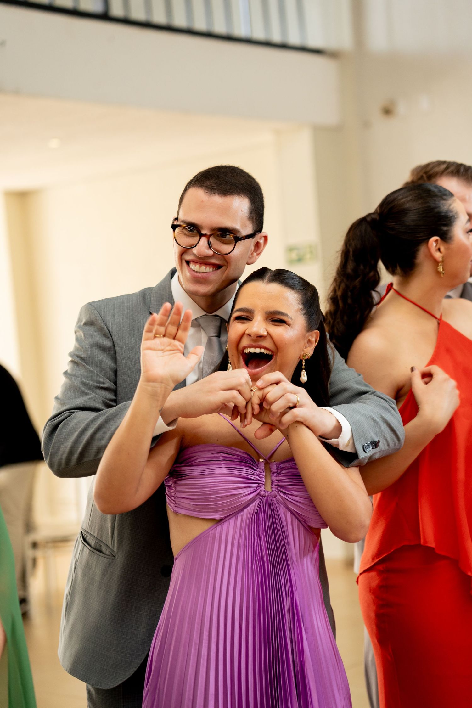 Couple dancing joyfully at a formal event, woman in purple dress making heart shape, man in gray suit smiling.