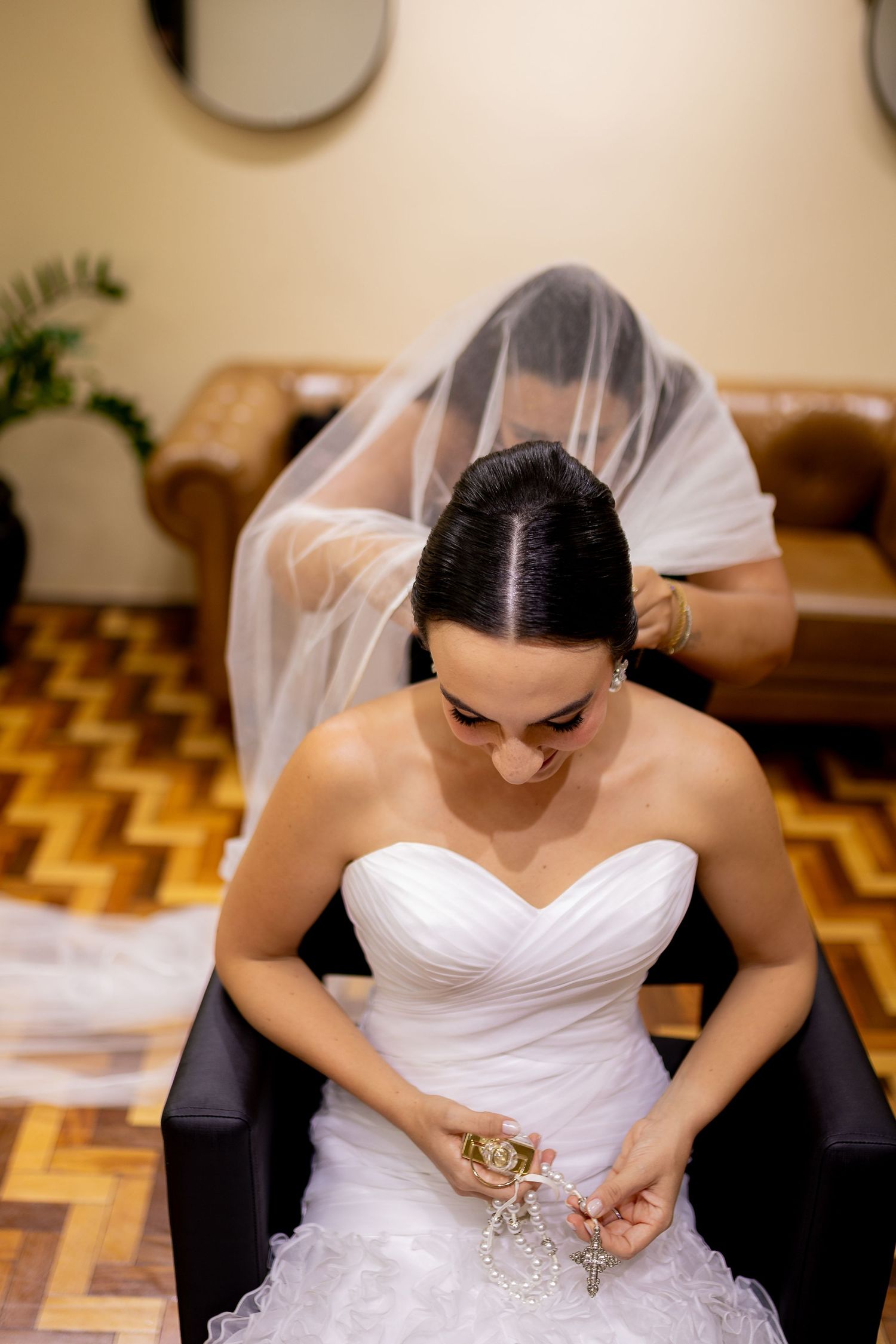Bride in white strapless gown examines pearl jewelry while bridesmaid adjusts veil during wedding preparations.