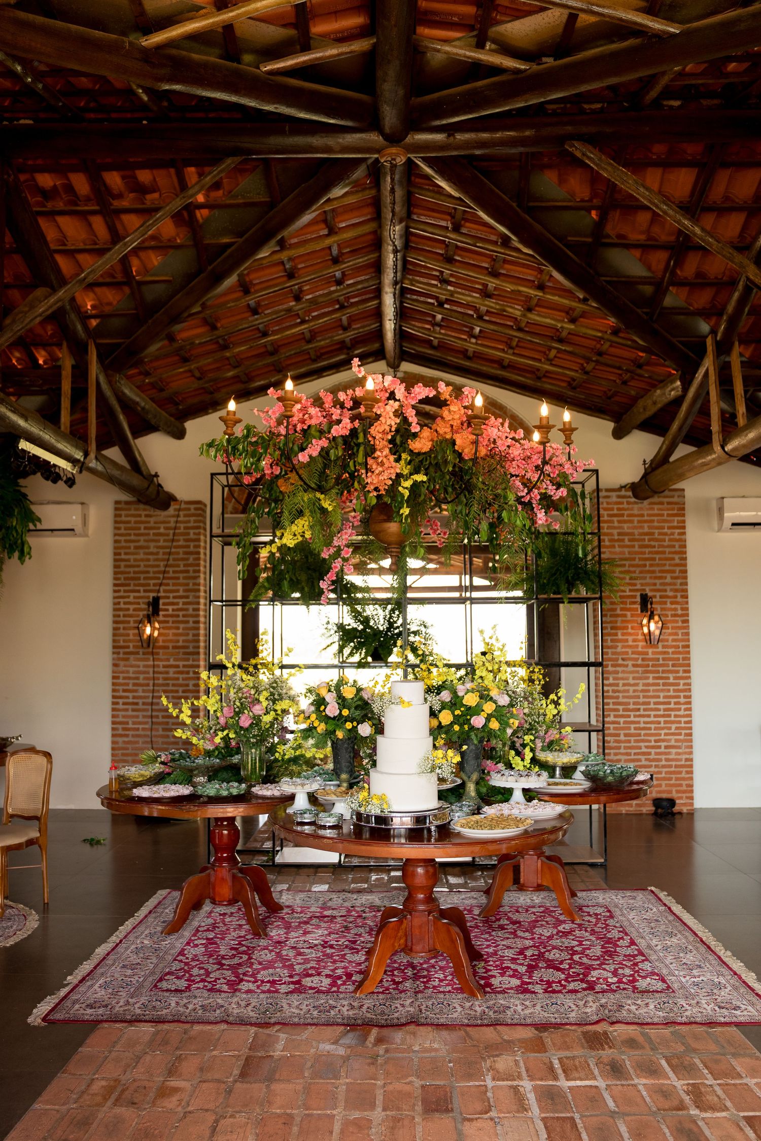 Elegant wedding cake table with pink floral chandelier, yellow blooms, and rustic brick venue with wooden ceiling.