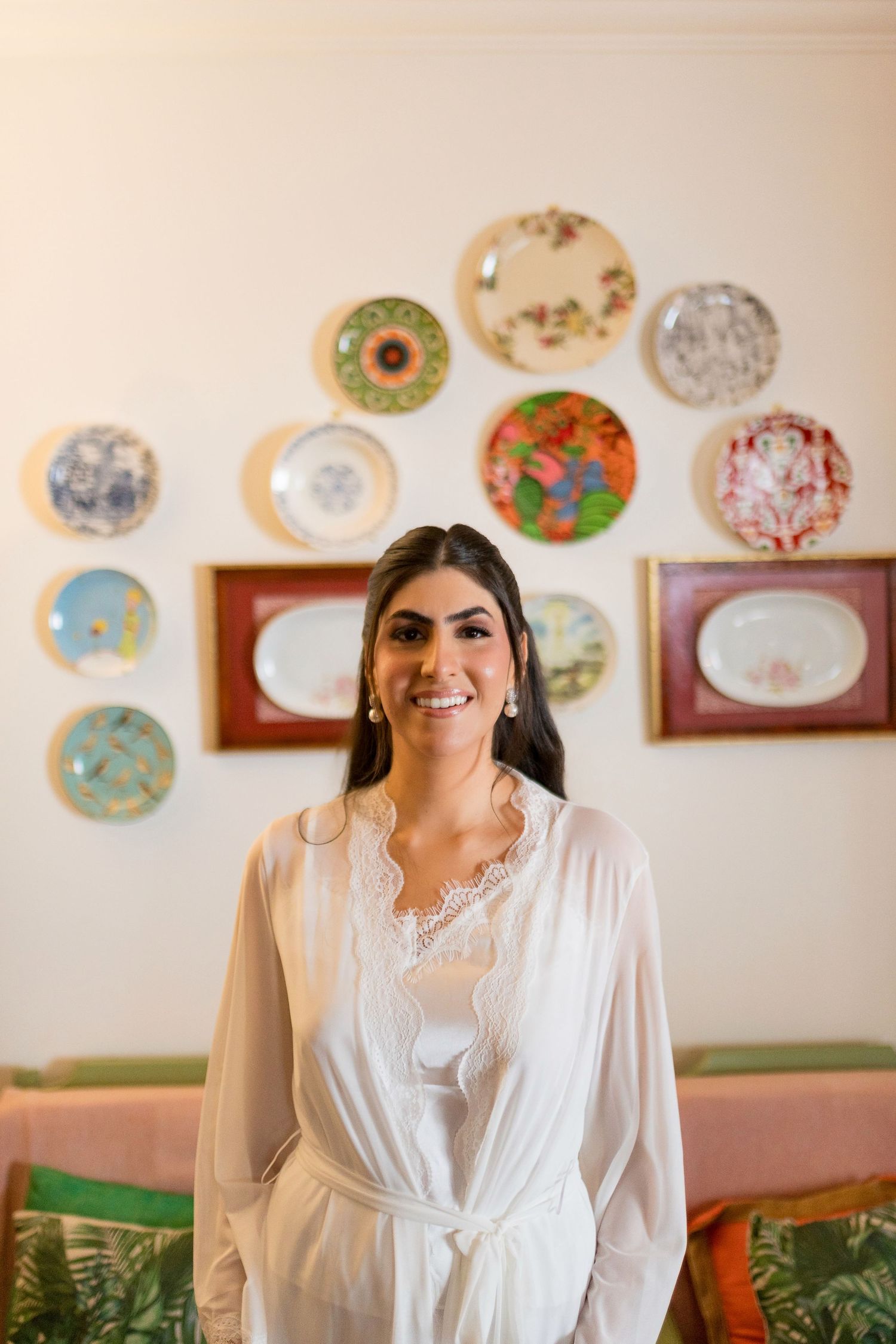 Smiling bride in white lace robe stands before a decorative wall display of colorful plates.