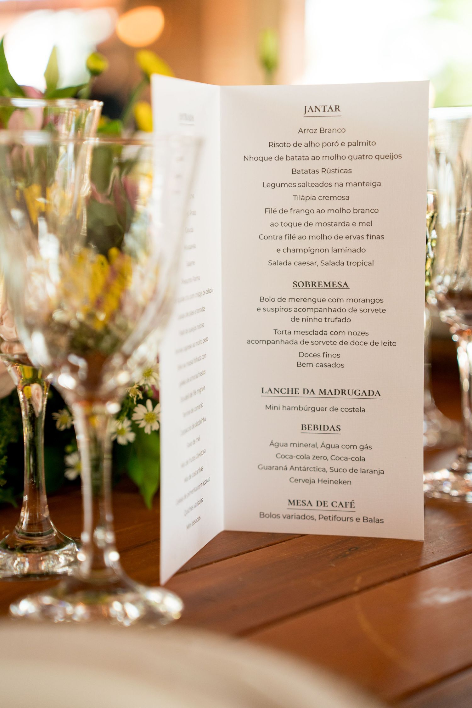 Elegant wedding reception menu card displayed upright on a table with crystal wine glasses and floral decor.