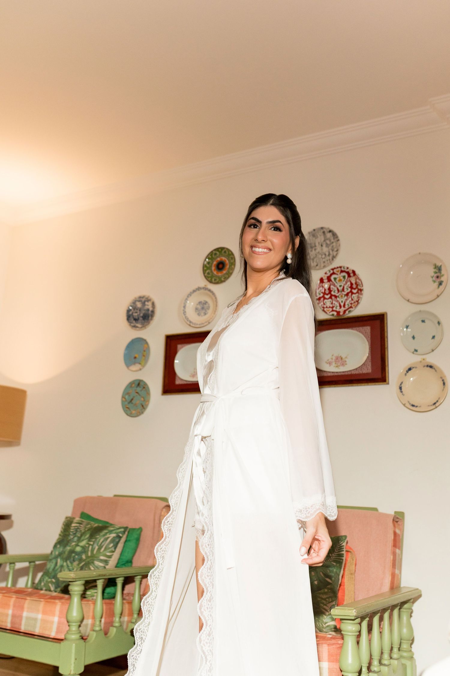Smiling bride in white lace robe poses in elegant room with decorative plates on wall.