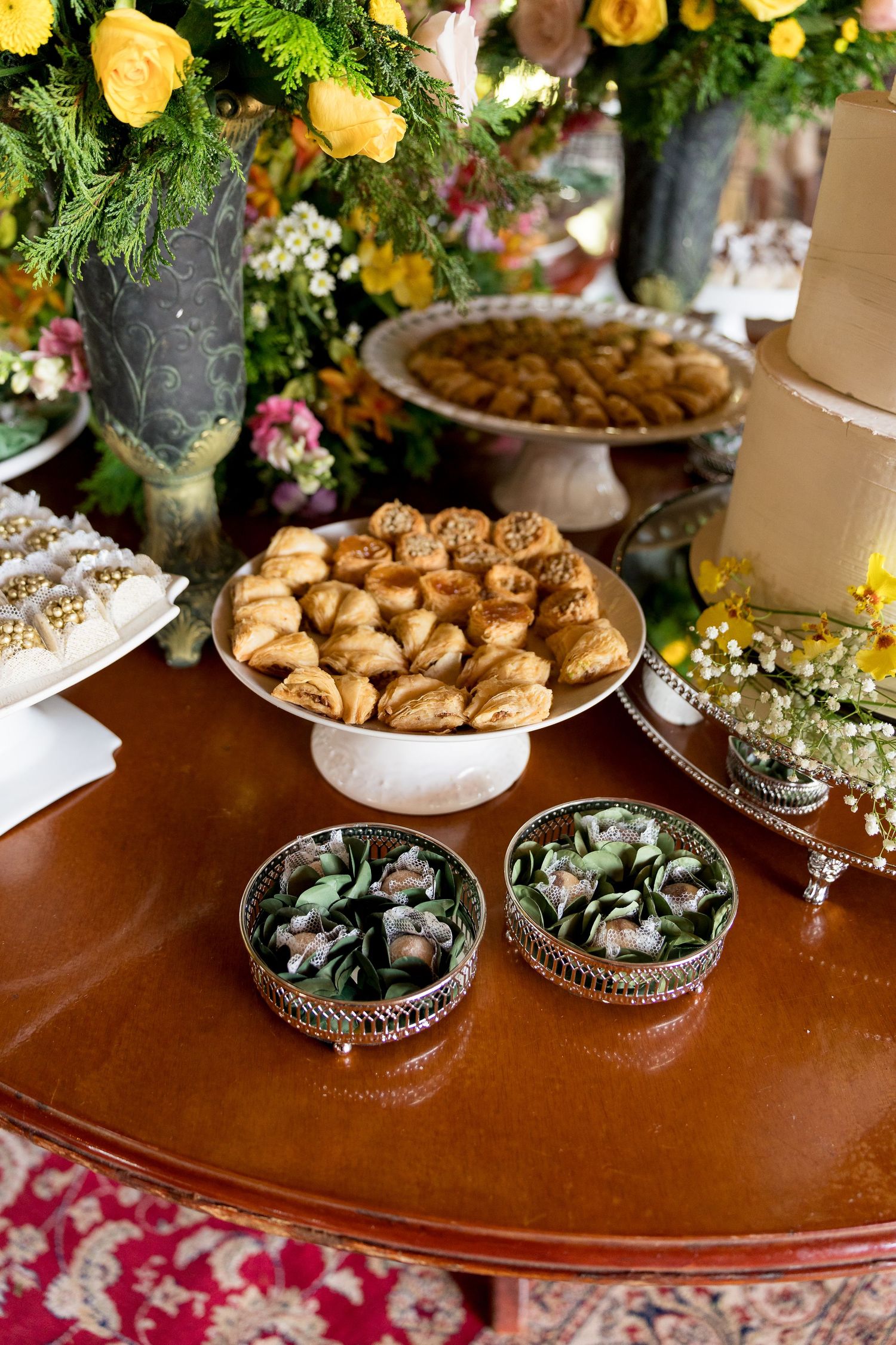 Elegant dessert table with assorted cookies on silver stands, wrapped candies, and yellow floral arrangements at a formal event.