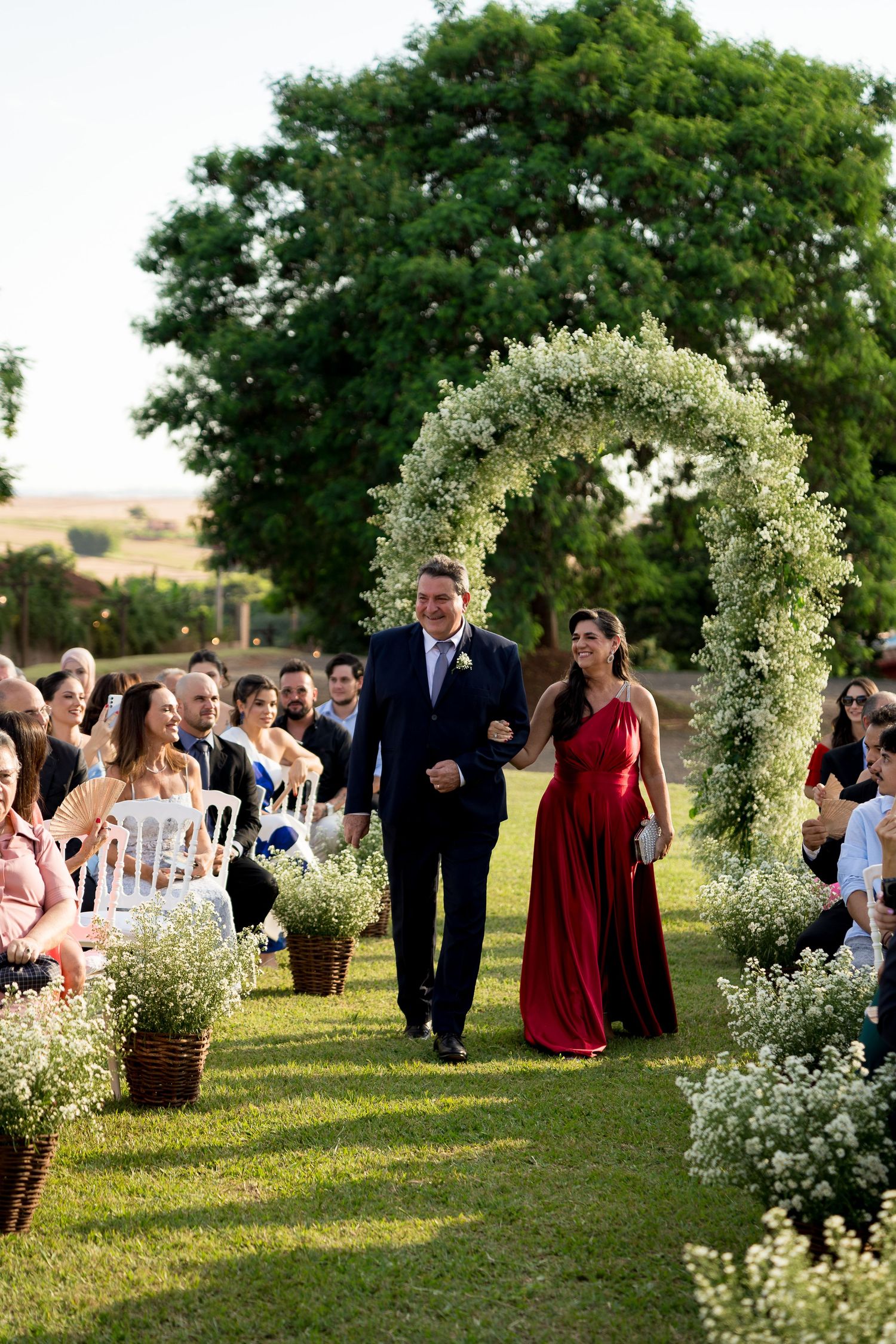 A couple walks down a floral arch-lined outdoor wedding aisle, guests seated on either side amid white flower arrangements.