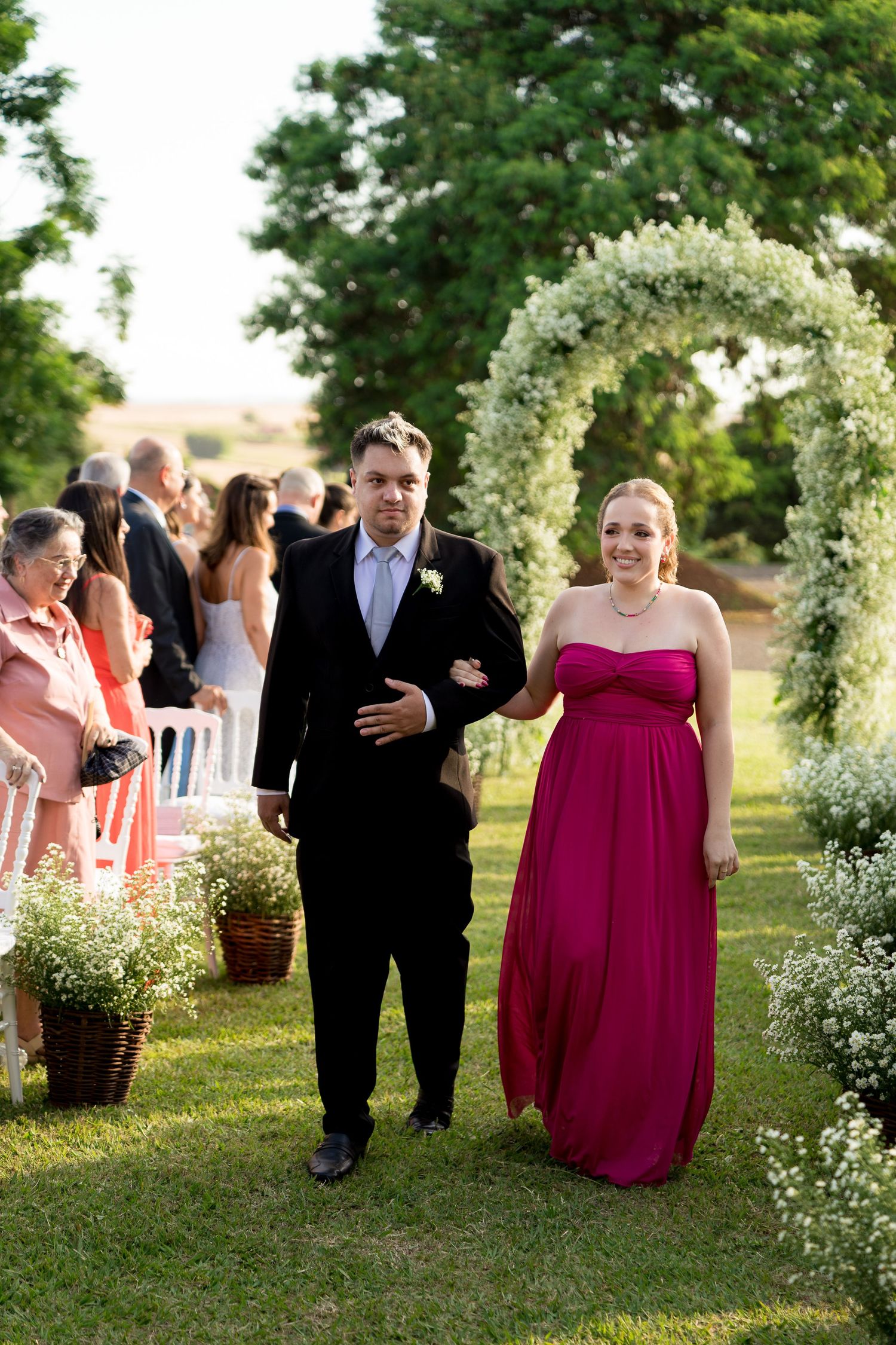 A groomsman in a black suit escorts a bridesmaid in a fuchsia gown down a floral-arched outdoor wedding aisle.