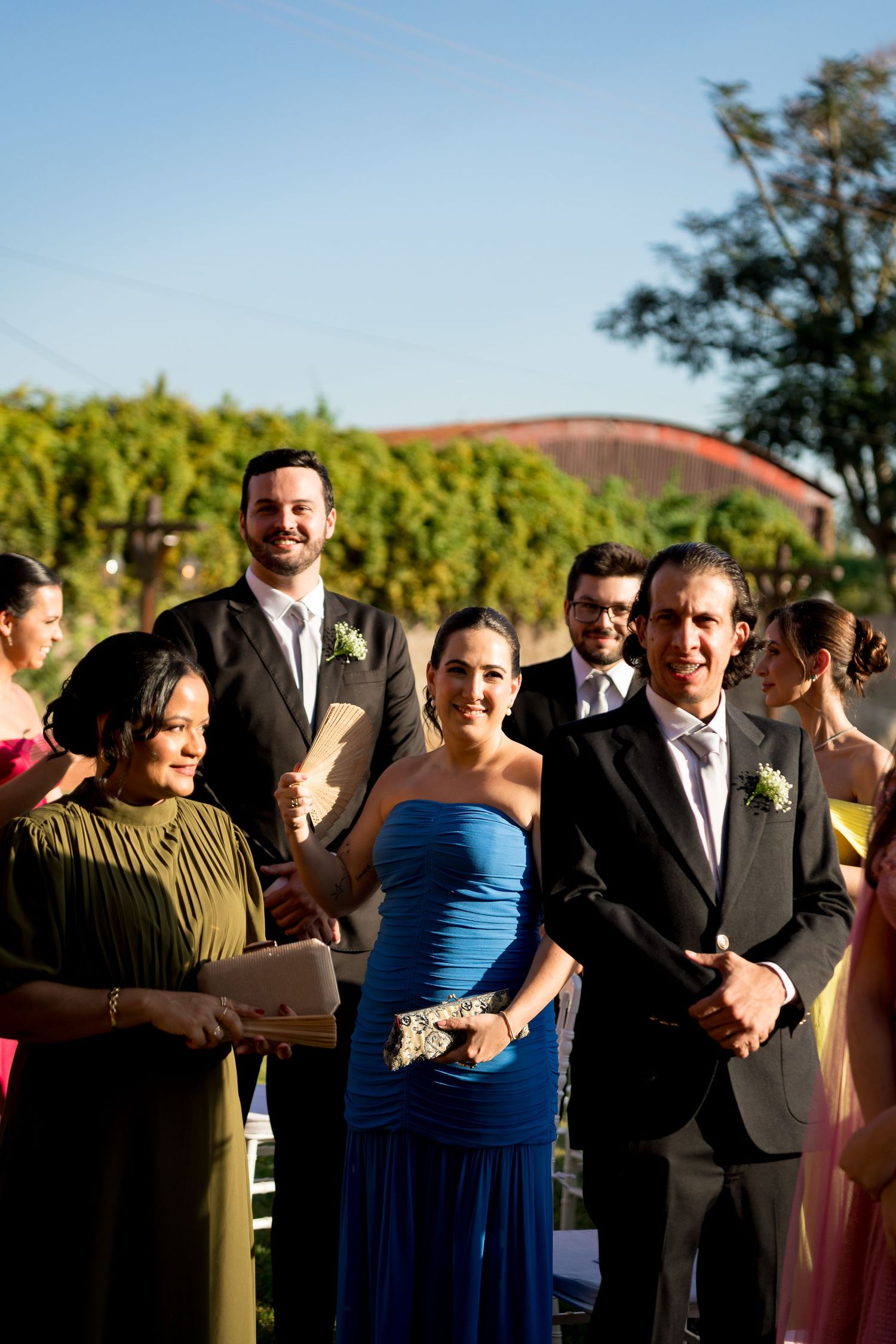Guests smile during an outdoor wedding ceremony, with a woman in a blue strapless gown and a man in a gray suit standing prominently.