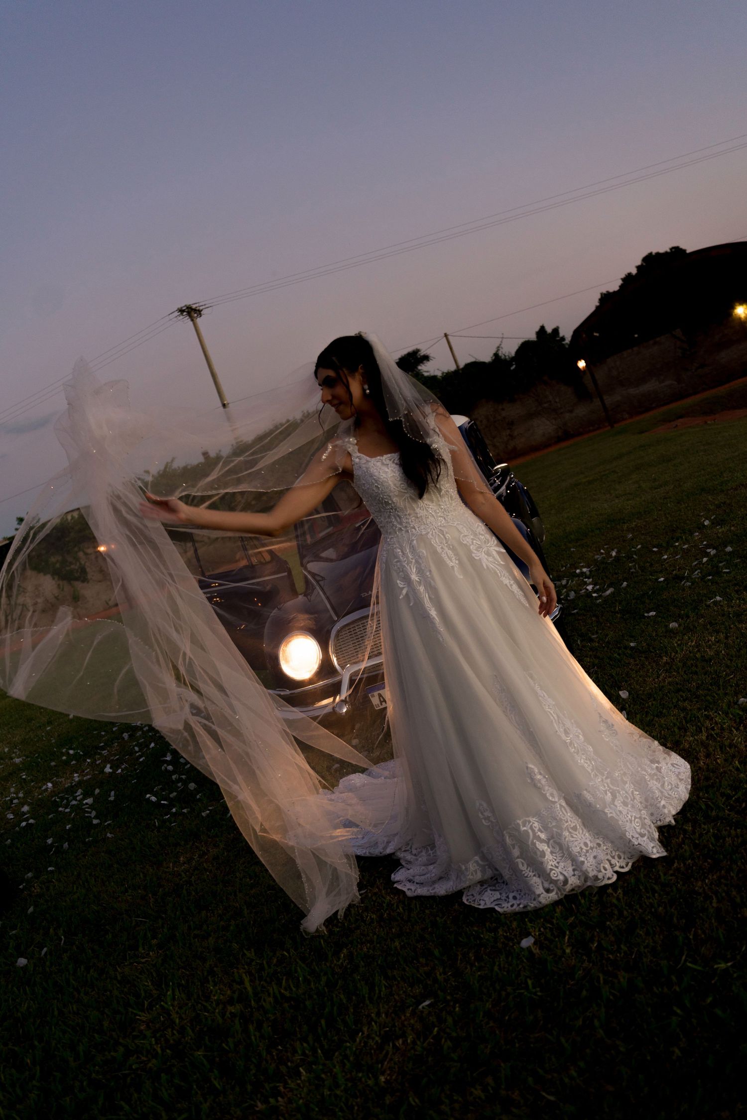 Bride in white lace ball gown with flowing veil poses outdoors at dusk near vintage car on green lawn.
