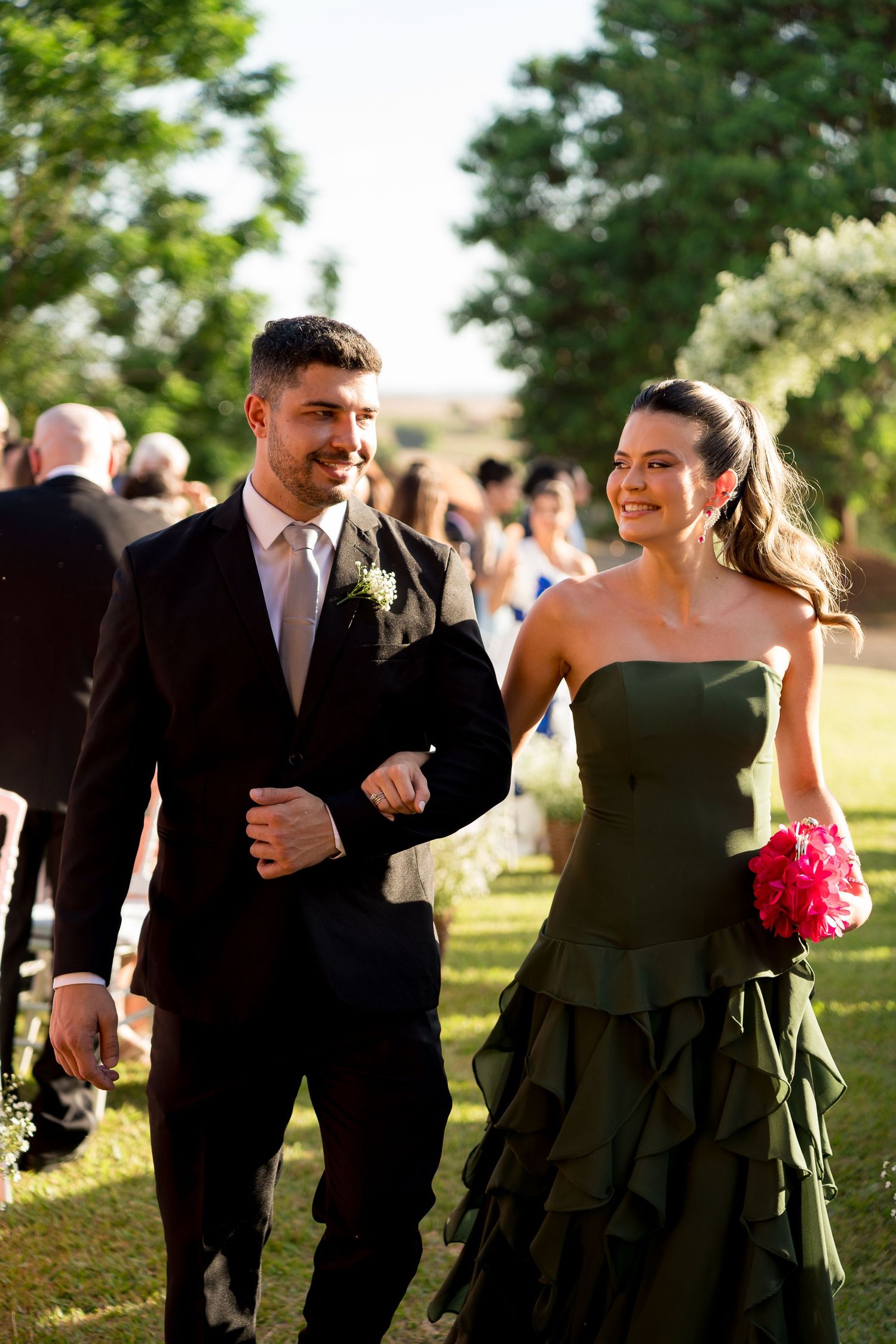 A smiling couple walks arm in arm at an outdoor wedding, man in dark suit, woman in strapless green gown holding pink flowers.