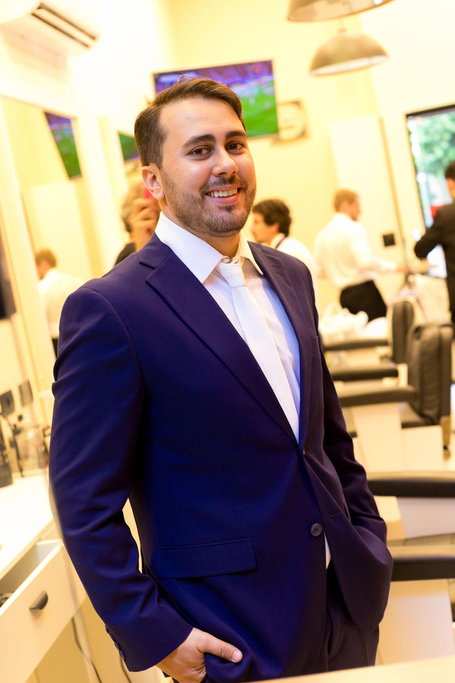 A smiling man in a navy blue suit stands confidently in a barbershop with TVs and customers visible in the background.