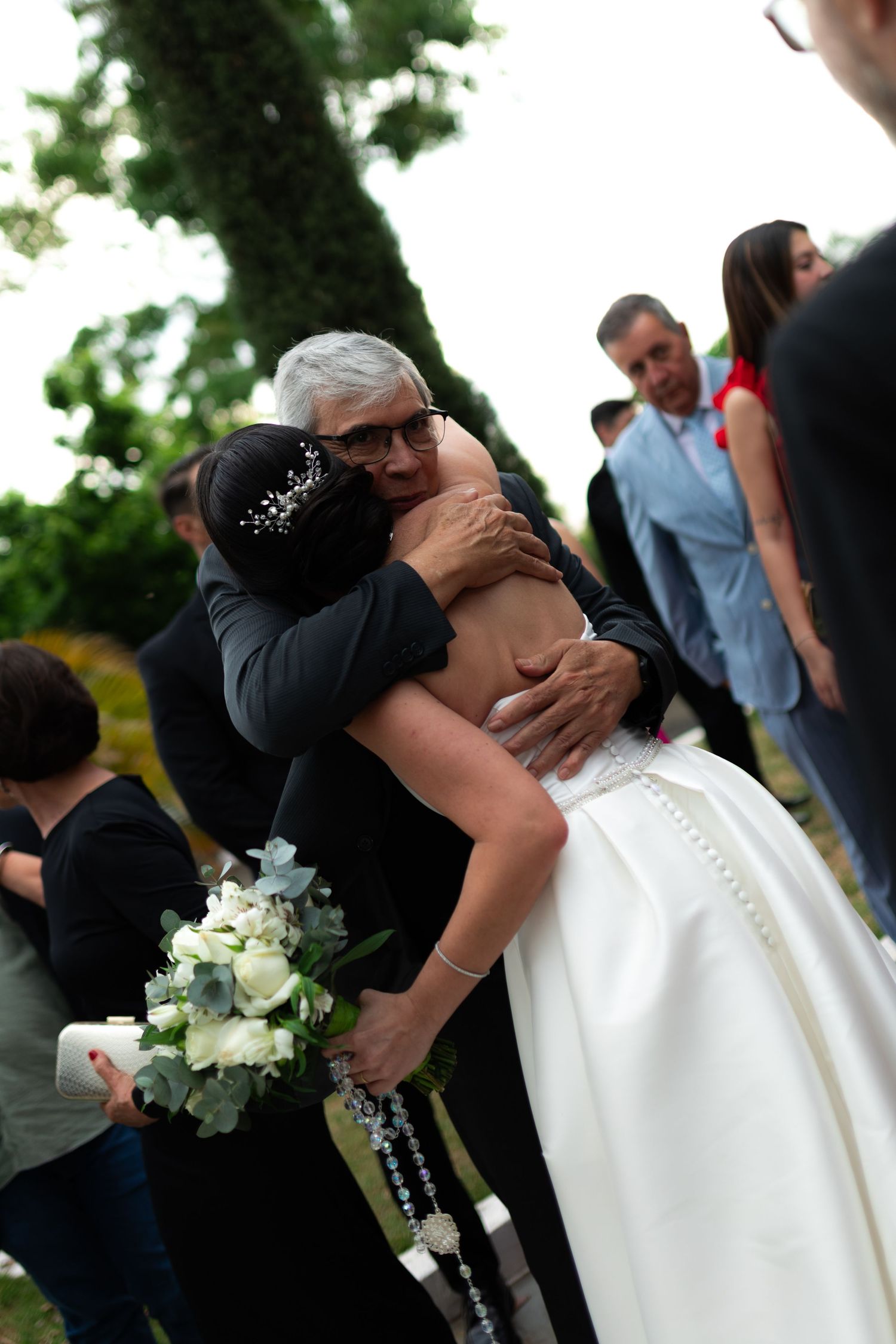 Bride in white gown embracing an older gentleman guest at outdoor wedding reception.