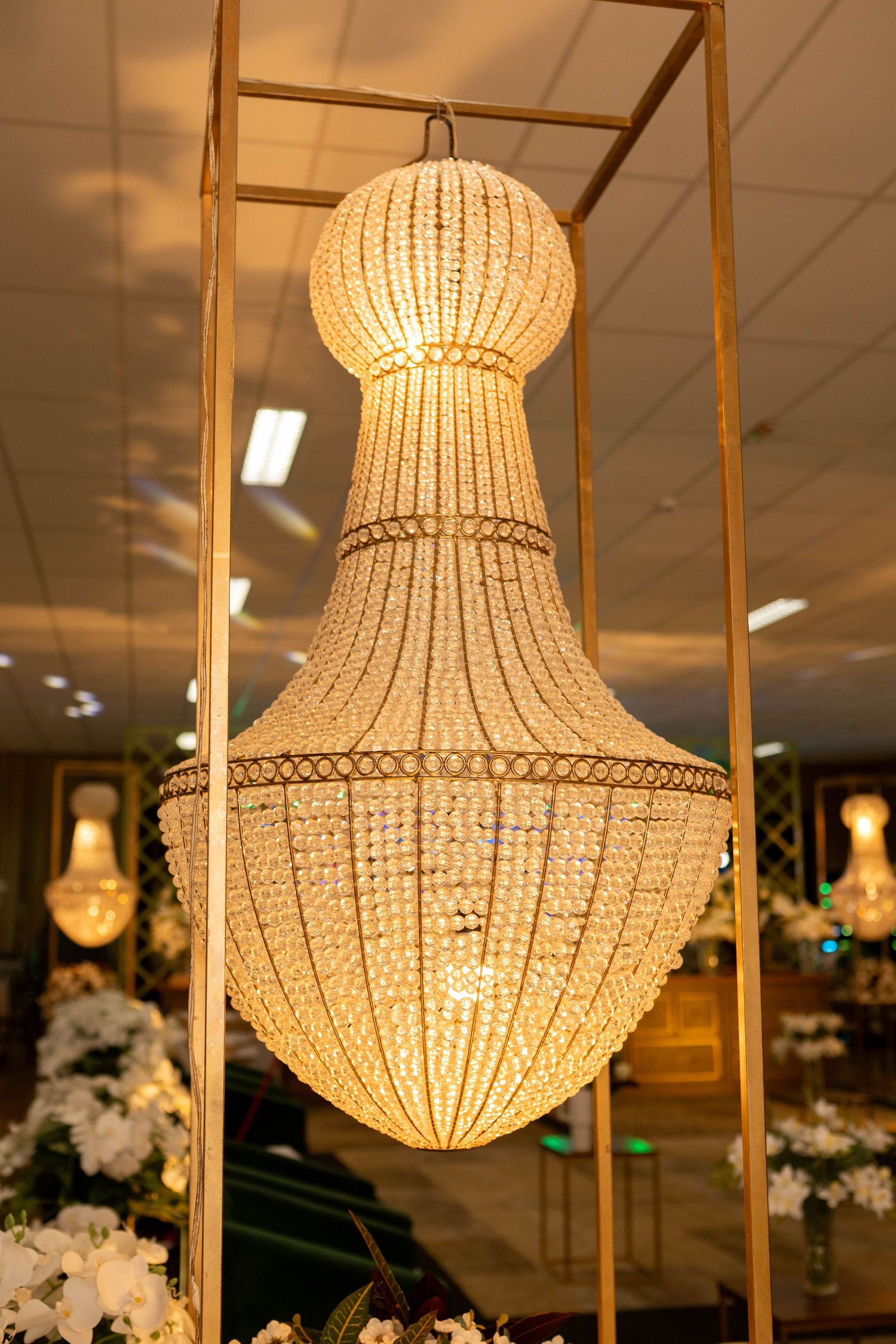 Elegant crystal chandelier with warm golden glow hanging in a decorated event space with white floral arrangements.