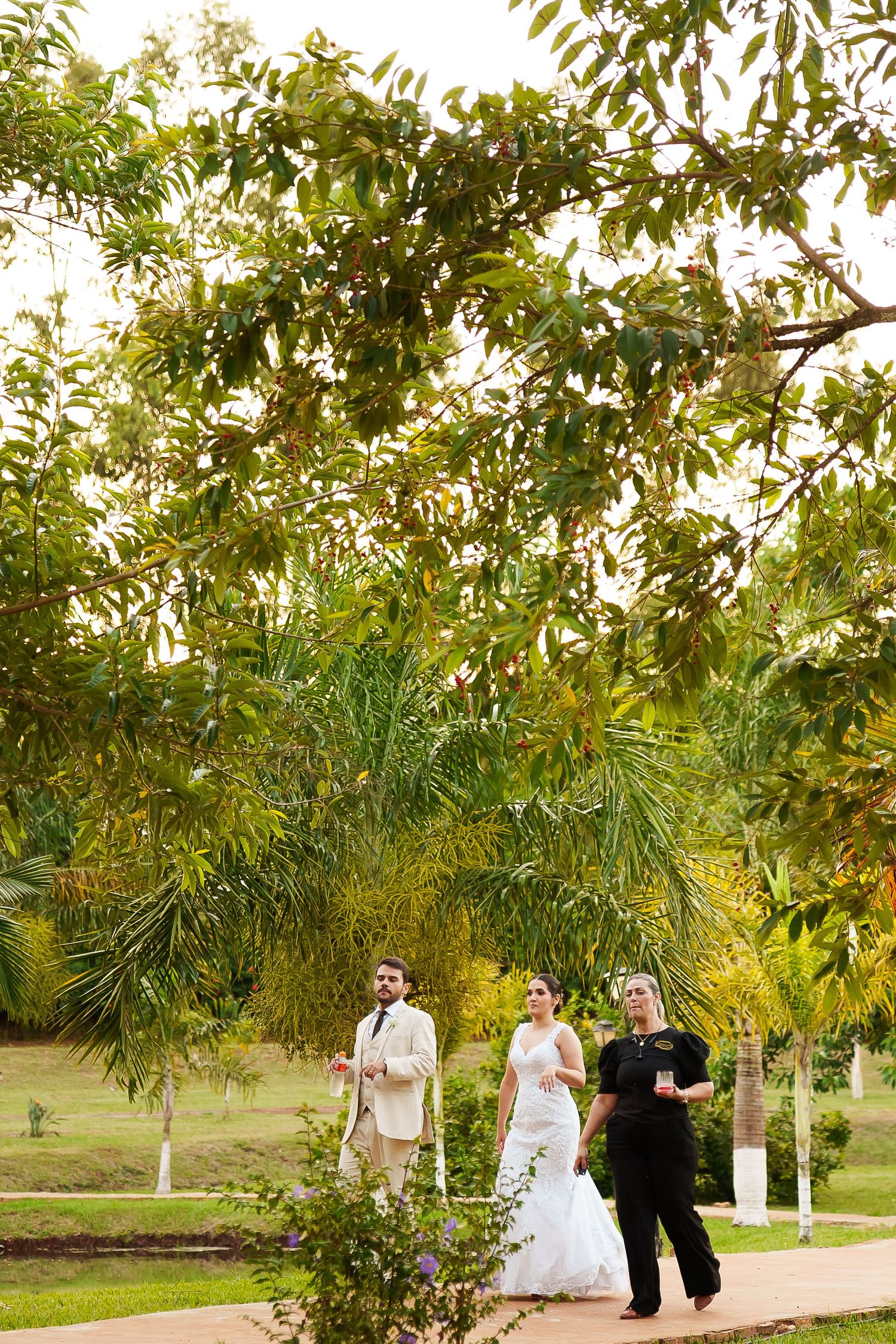 Bride in white gown and groom in tan suit walk with officiant through lush tropical garden on wedding day.