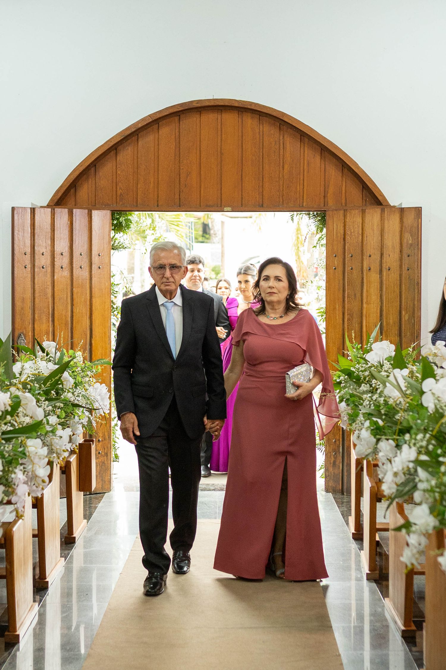 Elegant couple walking down a flower-adorned church aisle, man in dark suit, woman in mauve gown, wedding ceremony.