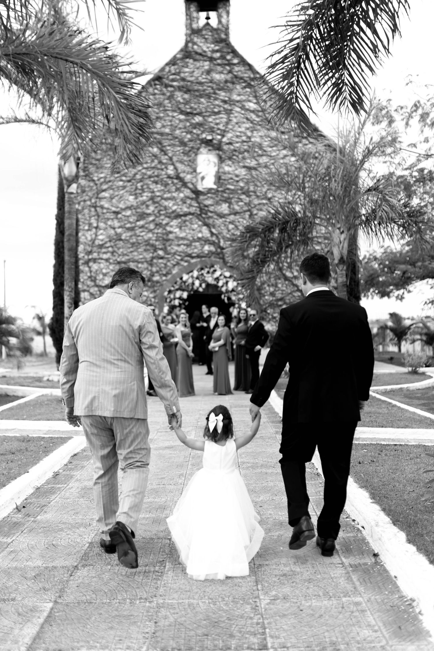 Black and white photo of flower girl walking hand in hand with two men toward a stone church wedding ceremony.