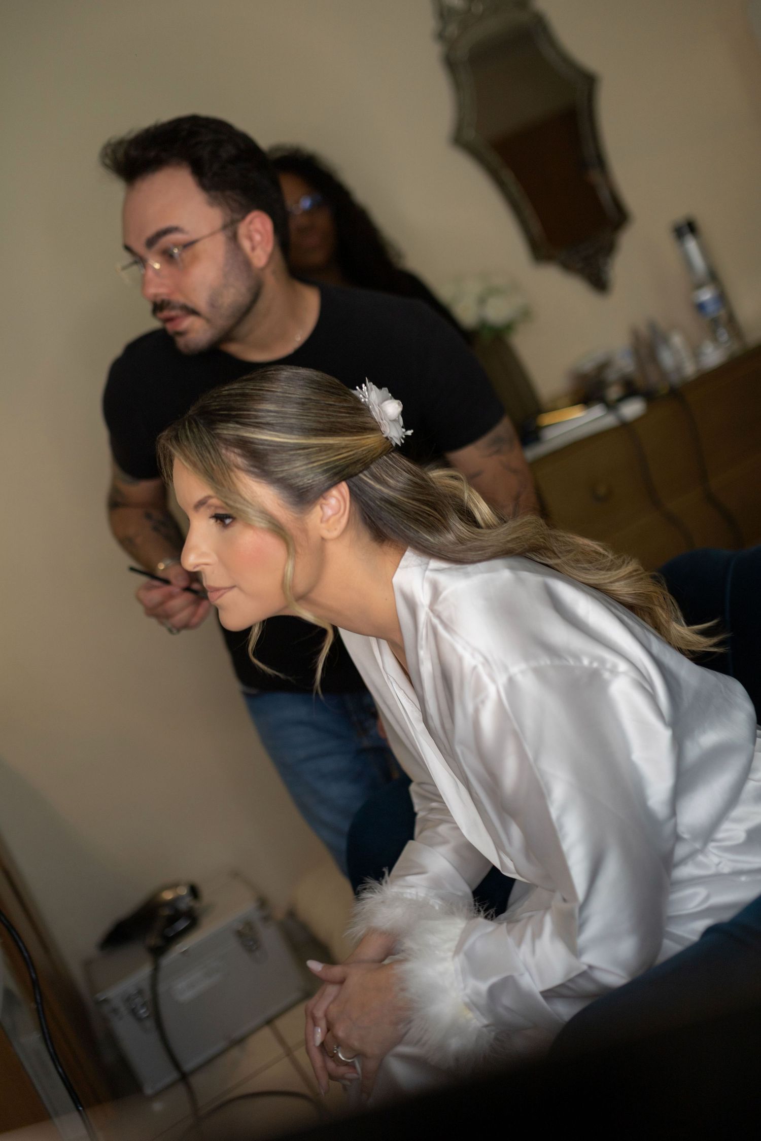 Bride in white satin robe gets makeup applied by artist before wedding ceremony.