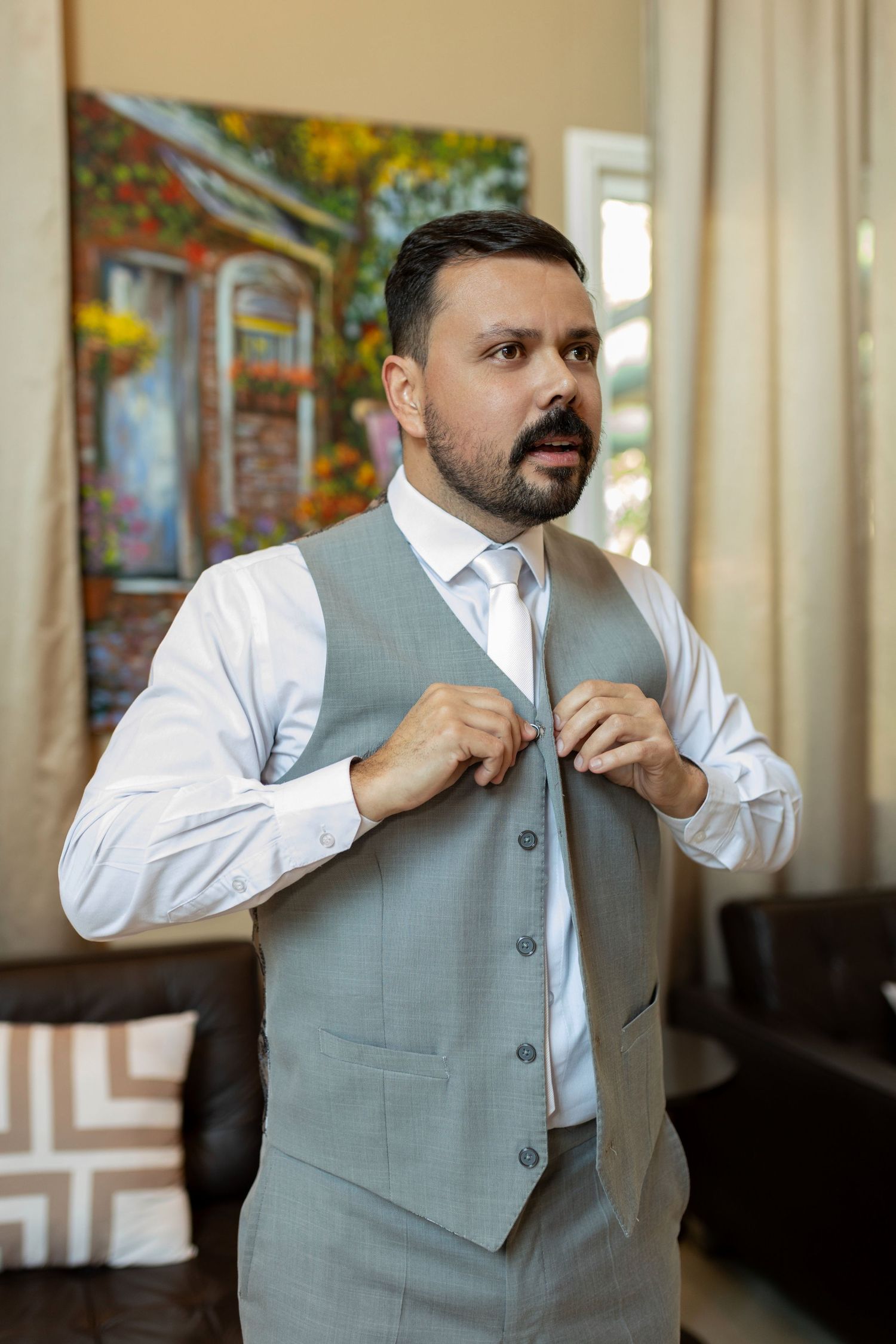 Man in gray vest and white shirt buttoning up while standing in elegant room with colorful painting.