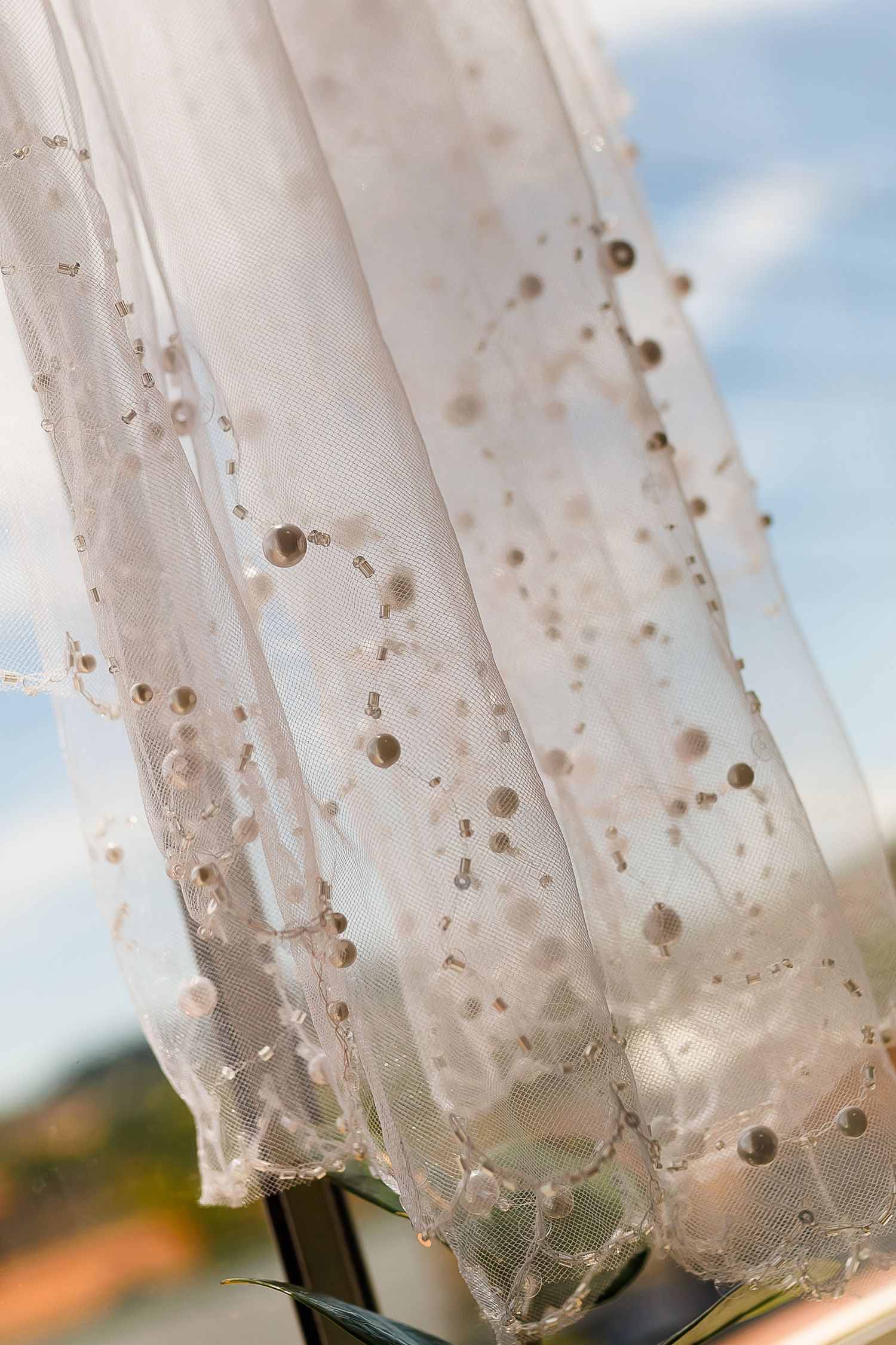 Delicate white lace wedding veil with pearl embellishments flowing against a blue sky backdrop.