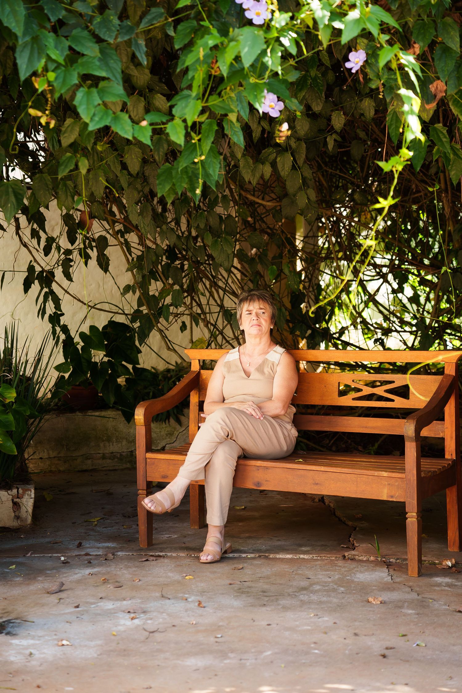 Woman in beige outfit sitting relaxed on wooden bench under lush green vine with purple flowers in sunlit garden.