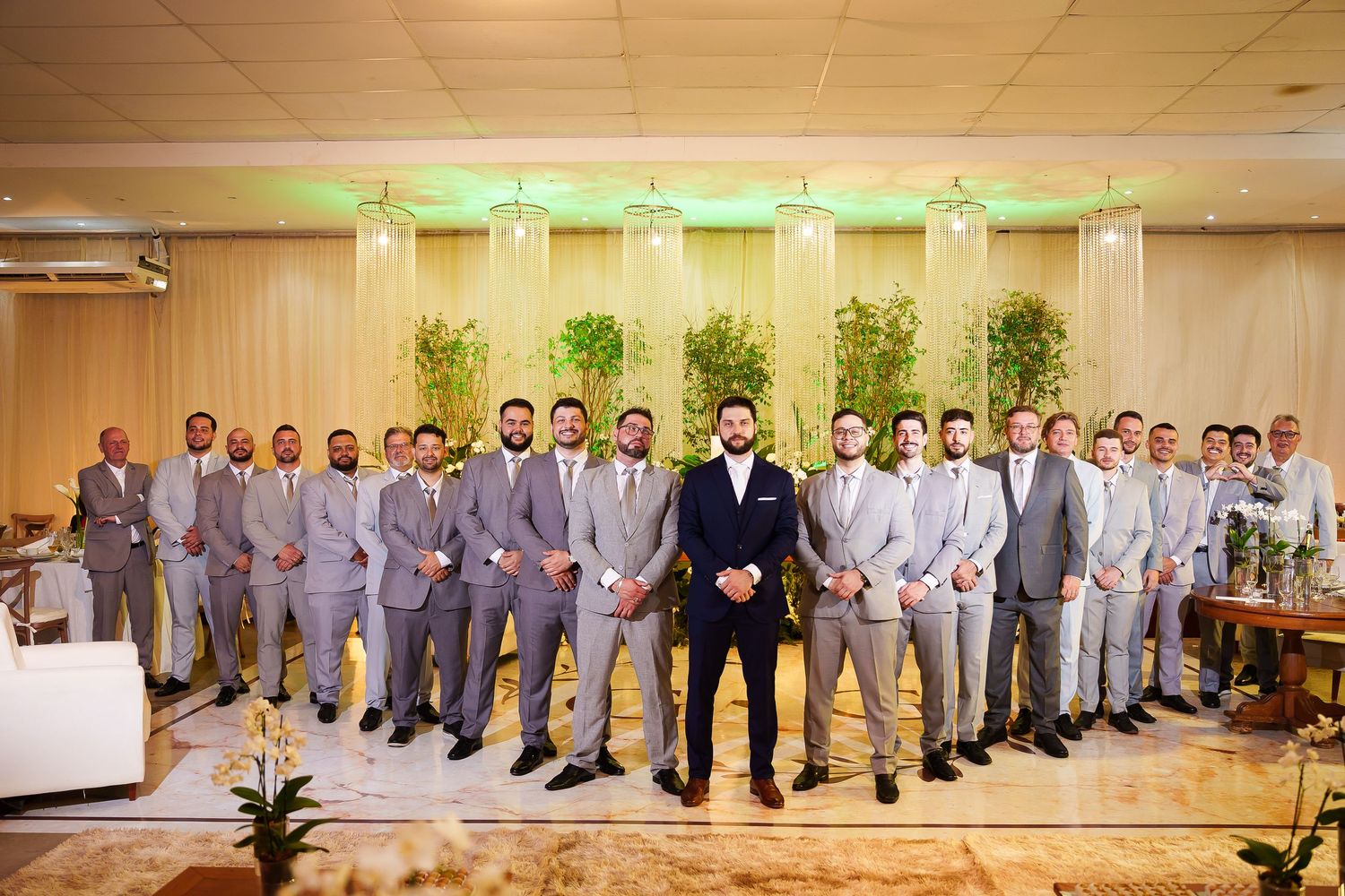 Groom in navy suit stands with groomsmen in gray suits at elegant wedding venue with green plants.